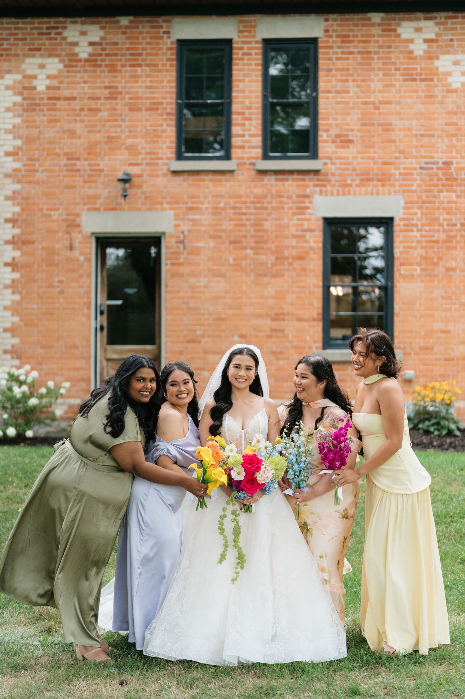 Wedding party in yellow dresses and olive suits laughs together in front of brick building.