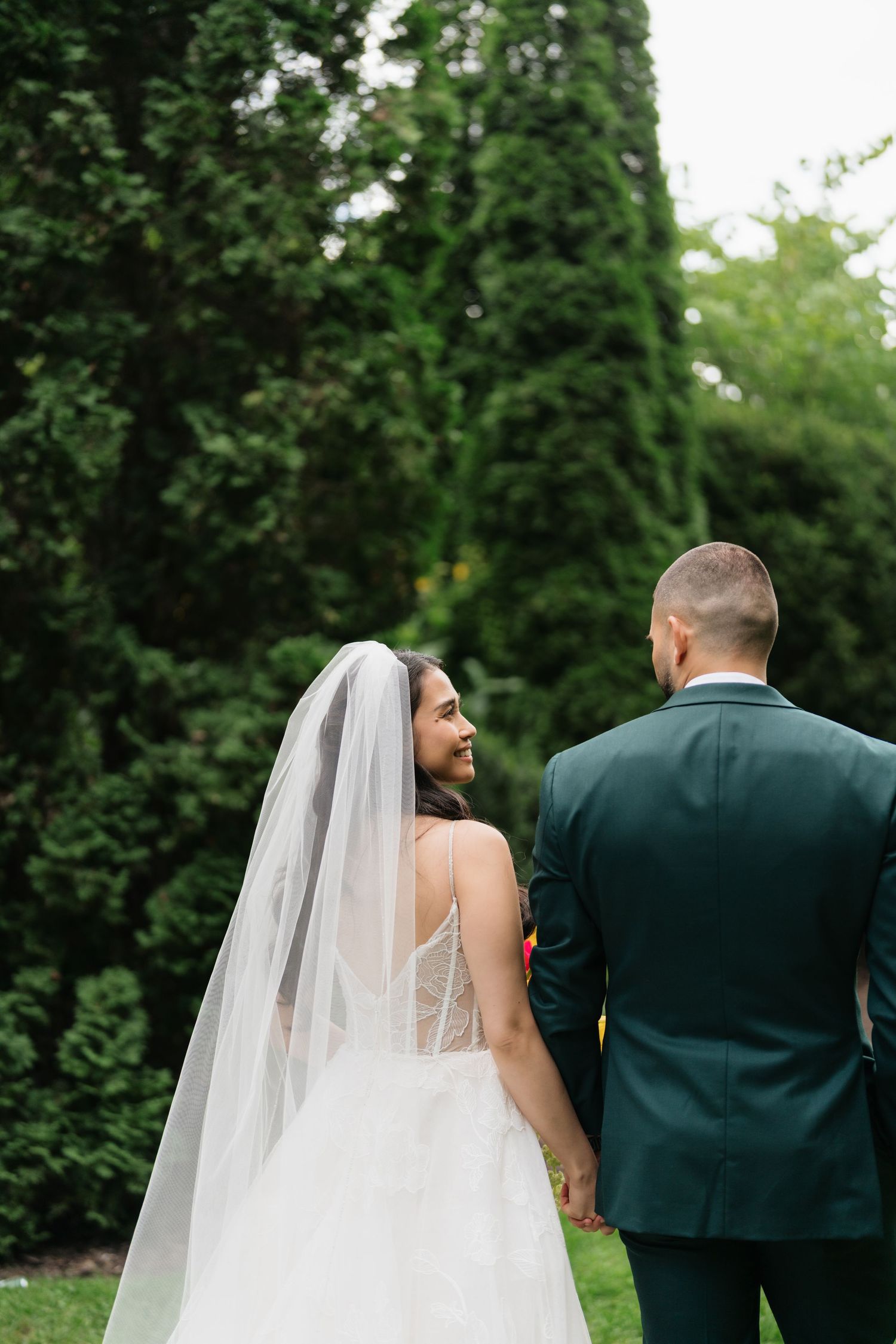 A couple in wedding attire stands together in a garden setting with lush green foliage in the background.