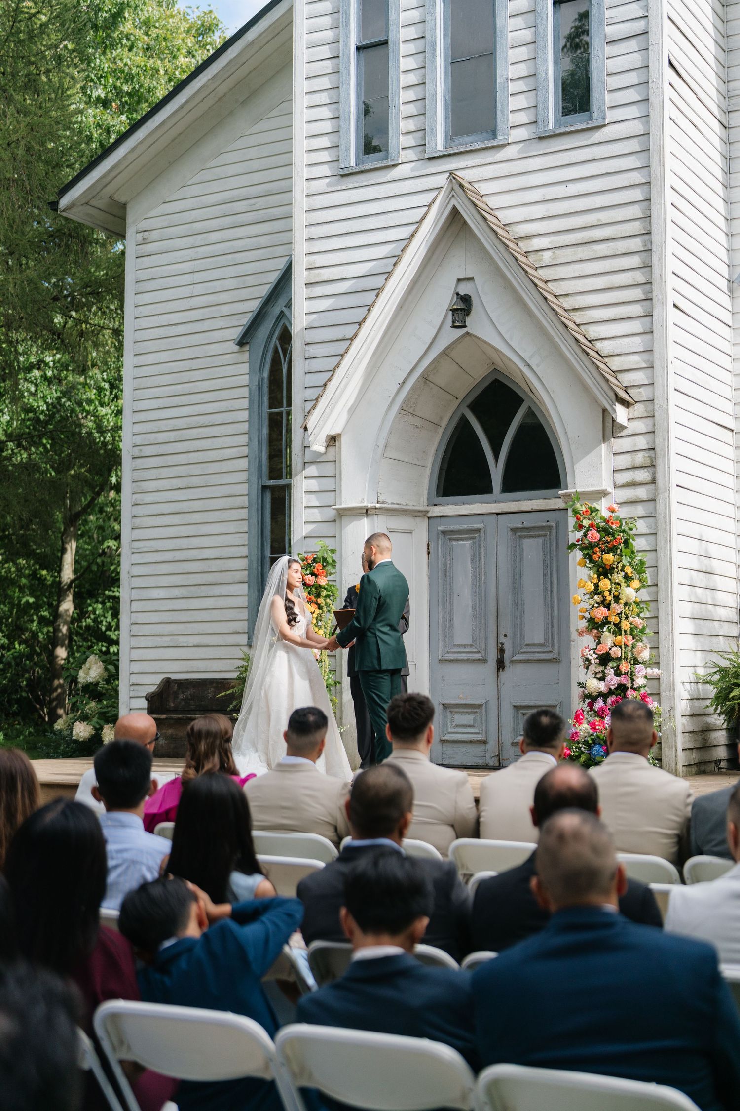 Wedding ceremony taking place at a charming white chapel with gothic arched doorway and floral decorations.