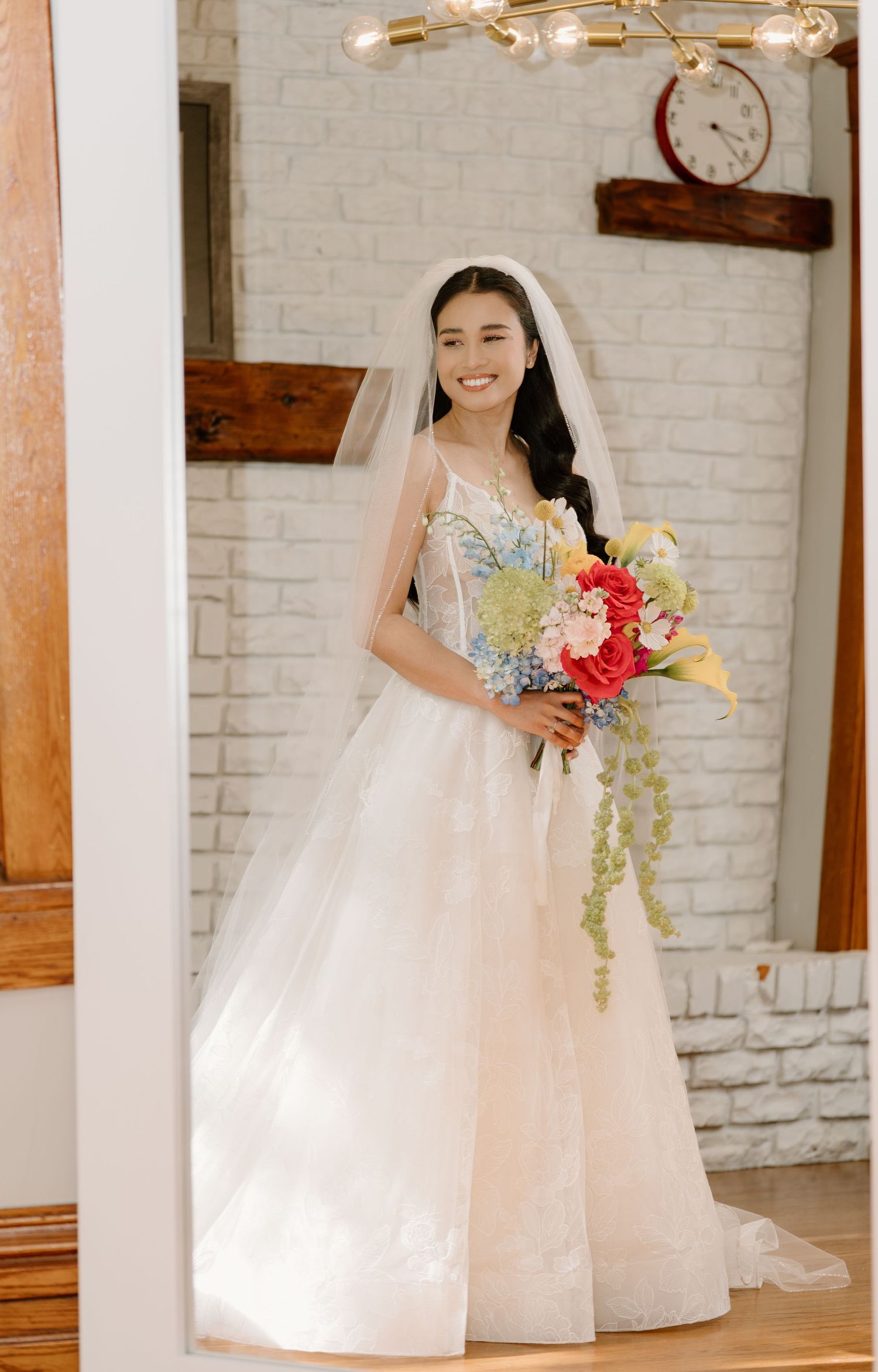 A person in a white wedding dress poses with a colorful floral bouquet in front of a white brick wall.