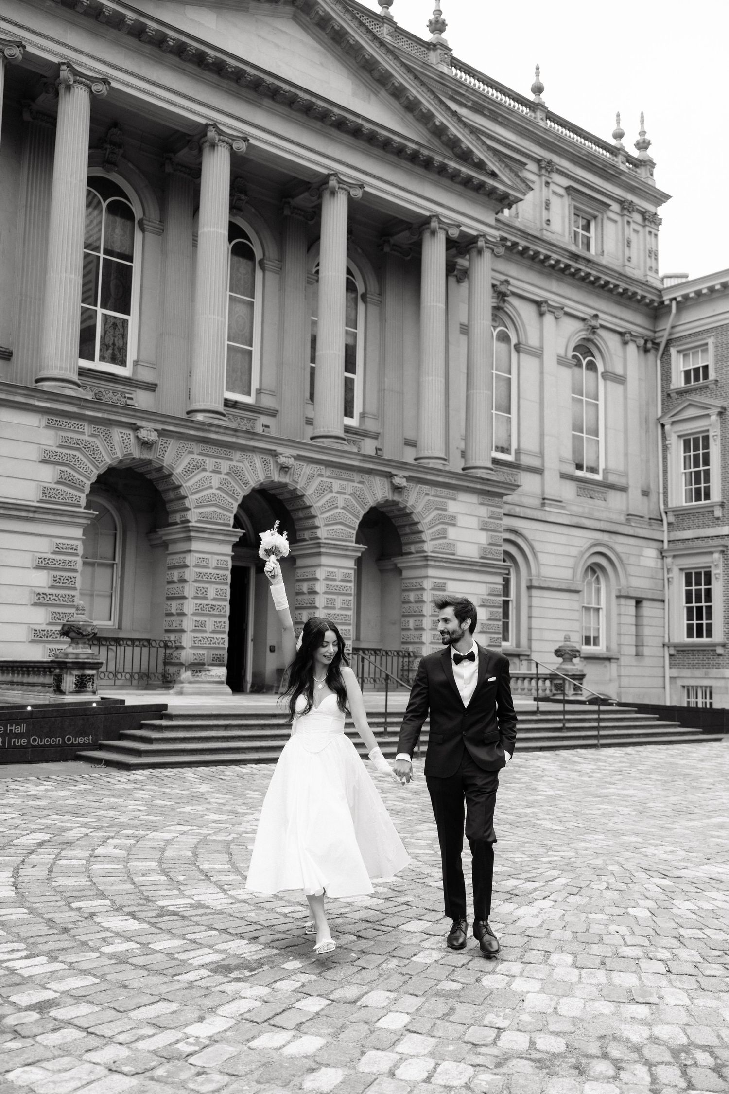 A newlywed couple walks hand in hand on cobblestone pavement in front of an elegant classical architecture building.
