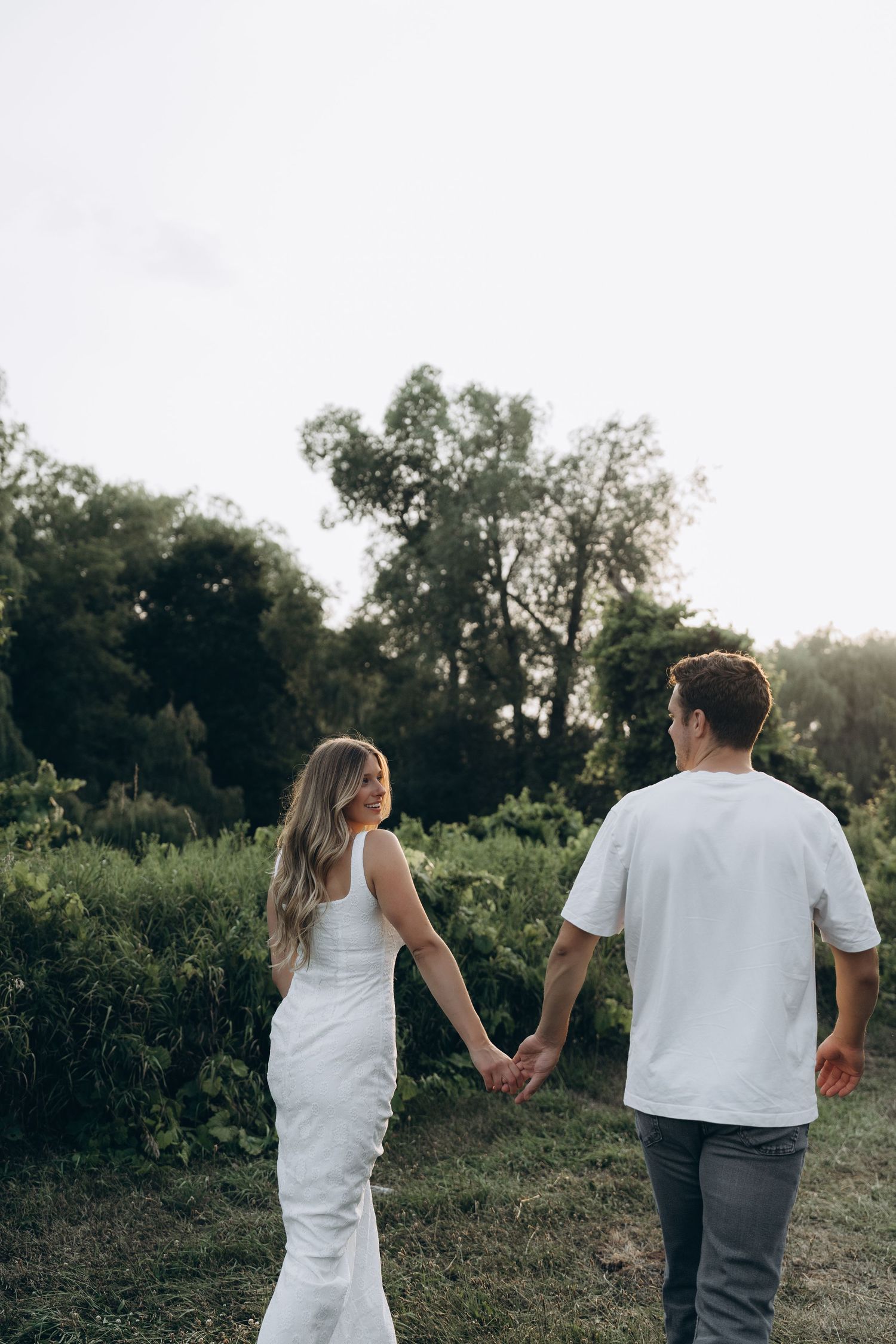 Two people in white outfits walk hand in hand through a natural outdoor setting with trees in background.