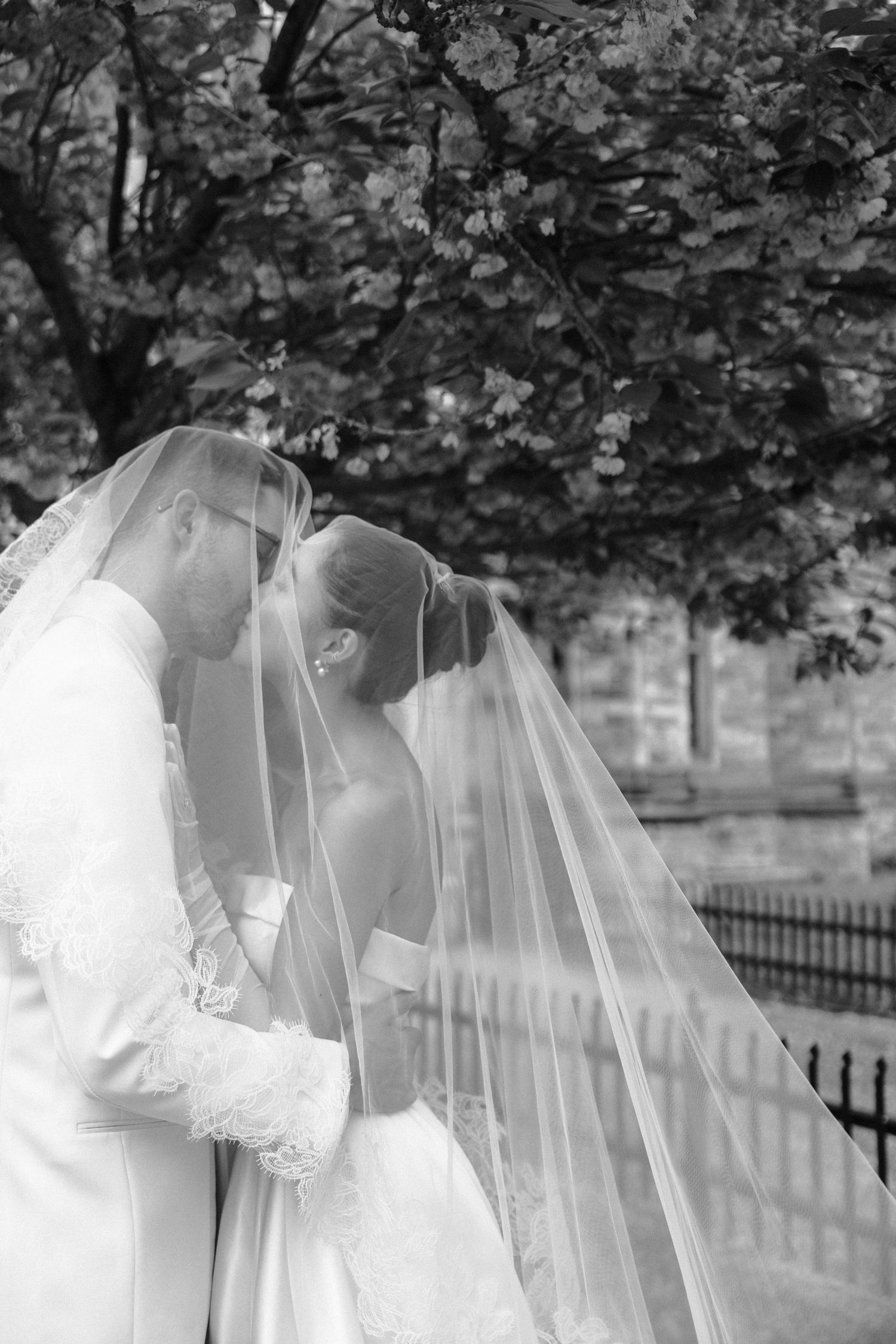 Two figures in elegant white wedding gowns and veils stand side by side in a romantic black and white outdoor scene.