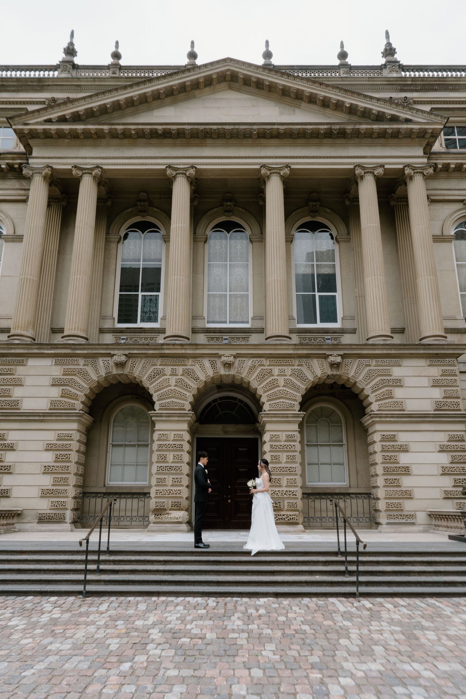 A wedding photo of a couple on the steps of a grand historic building with columns and arched windows.