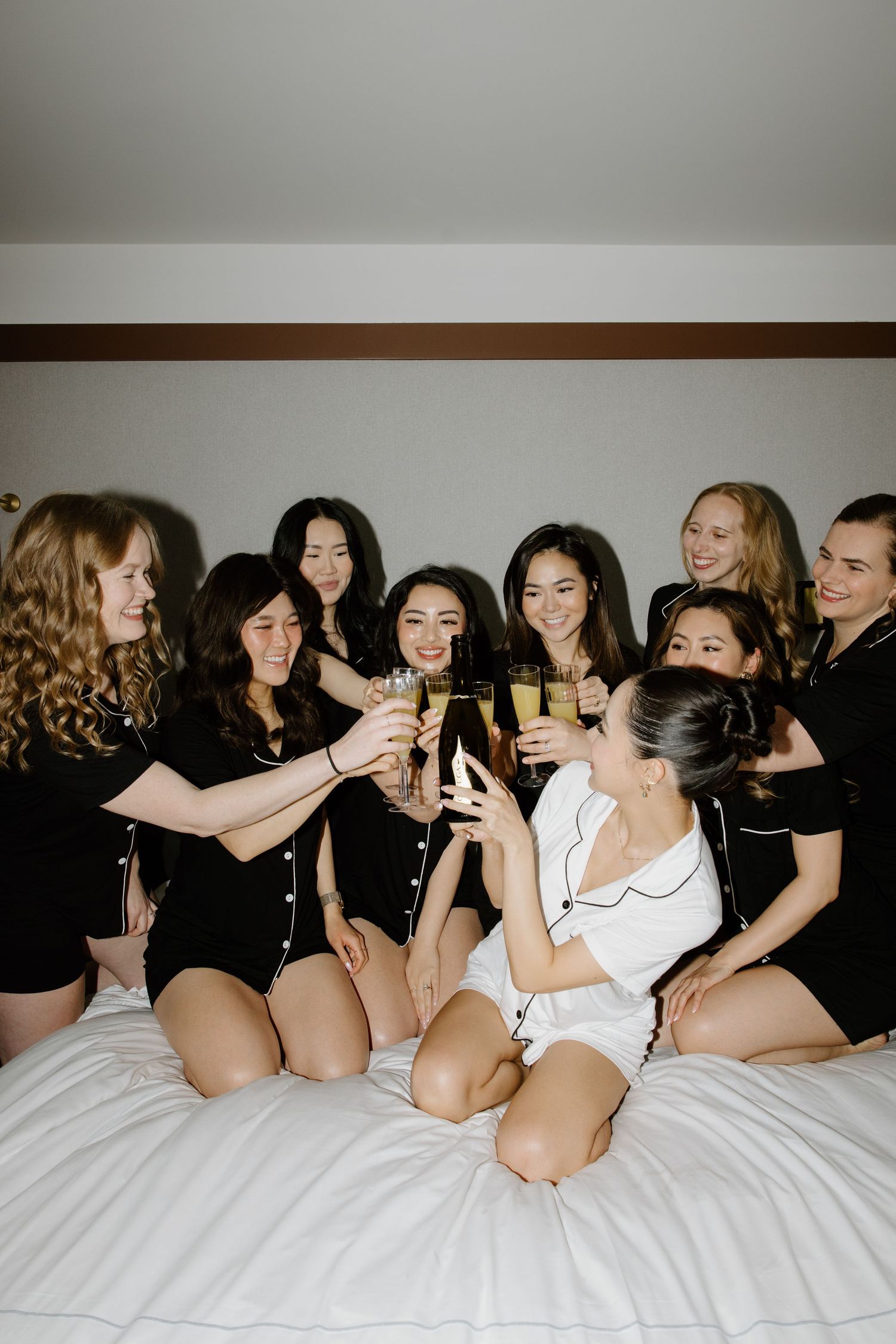 A group of friends in black and white outfits toasting with drinks while sitting together on a bed.