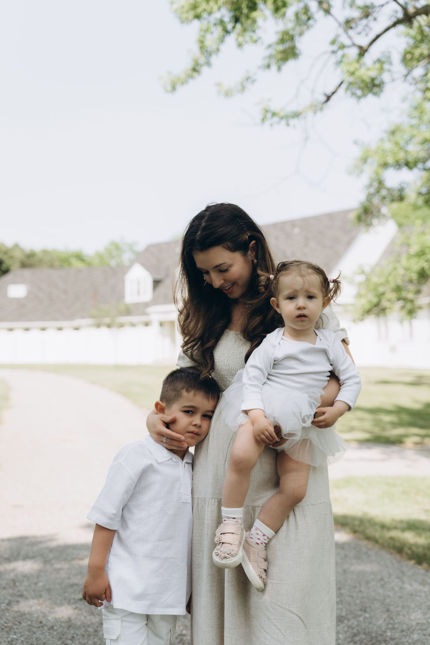 A family in coordinated neutral outfits poses together outdoors on a sunny day against a white building and trees.