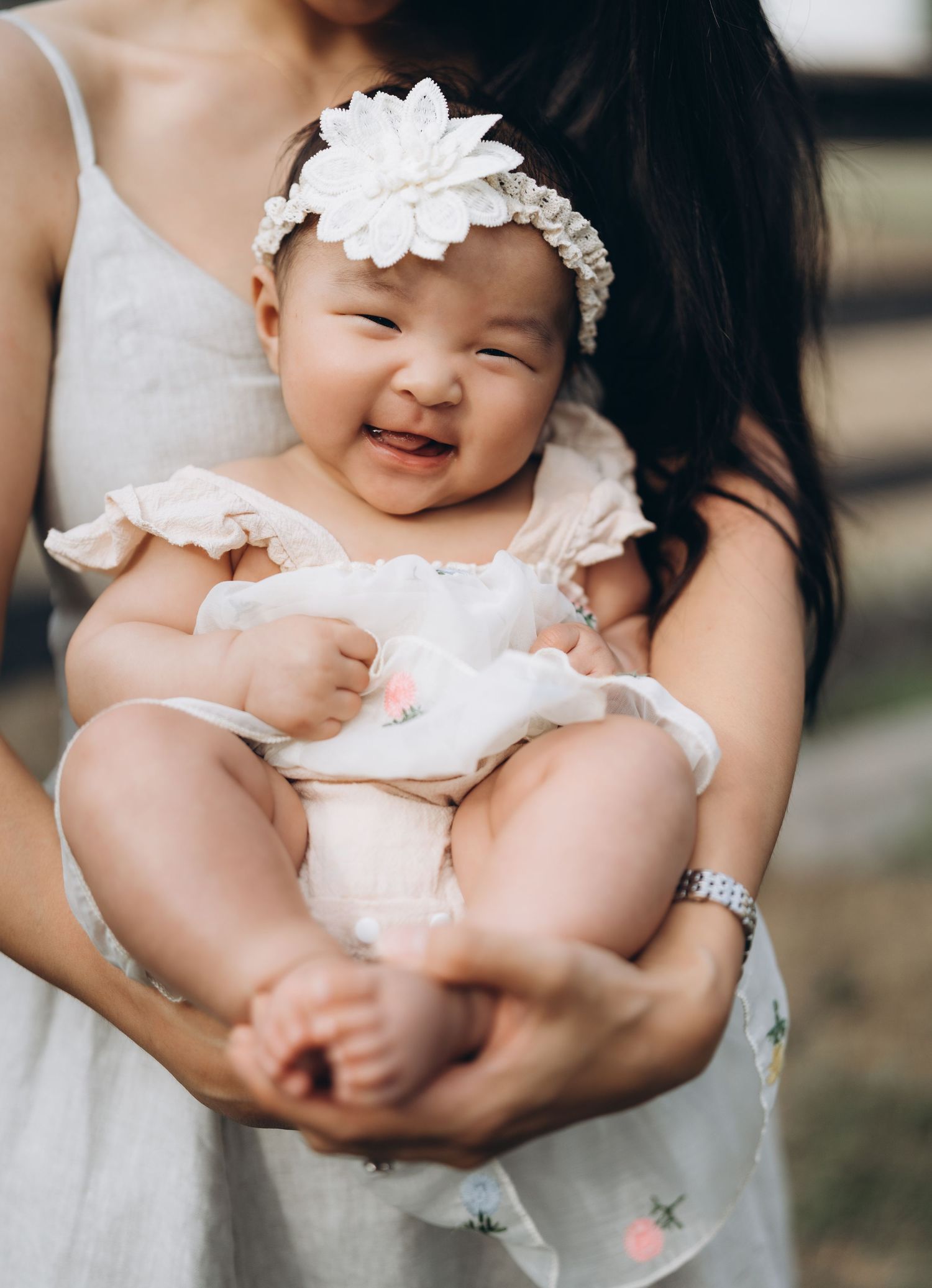A joyful baby wearing a white dress and floral headband smiles while being held by someone in white.