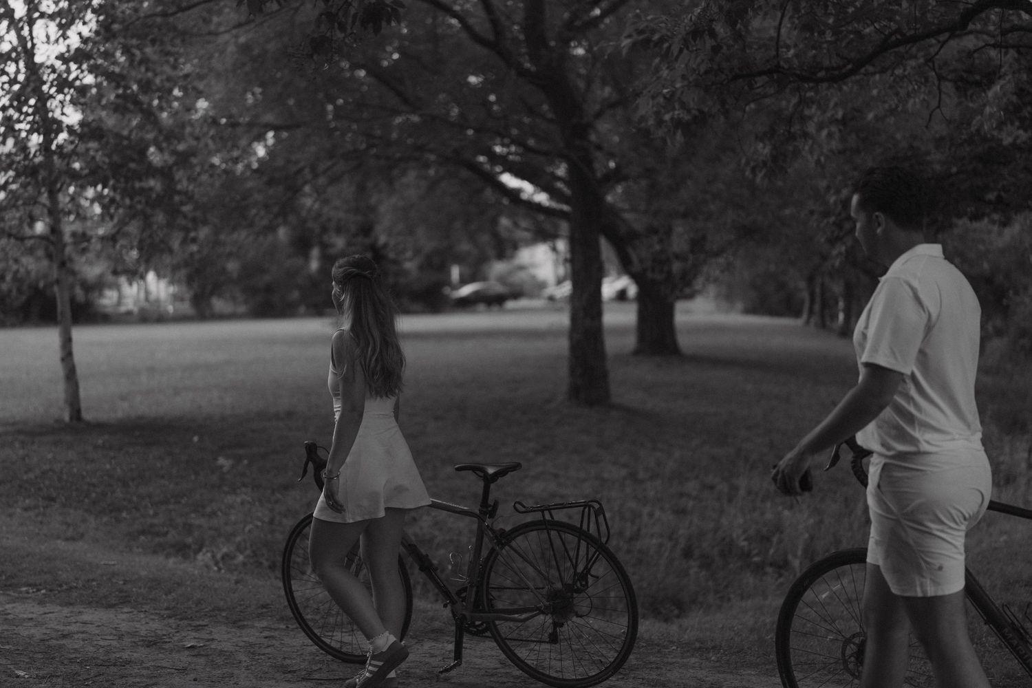 A black and white photograph of a figure sitting under a tree next to a parked bicycle in a park setting.