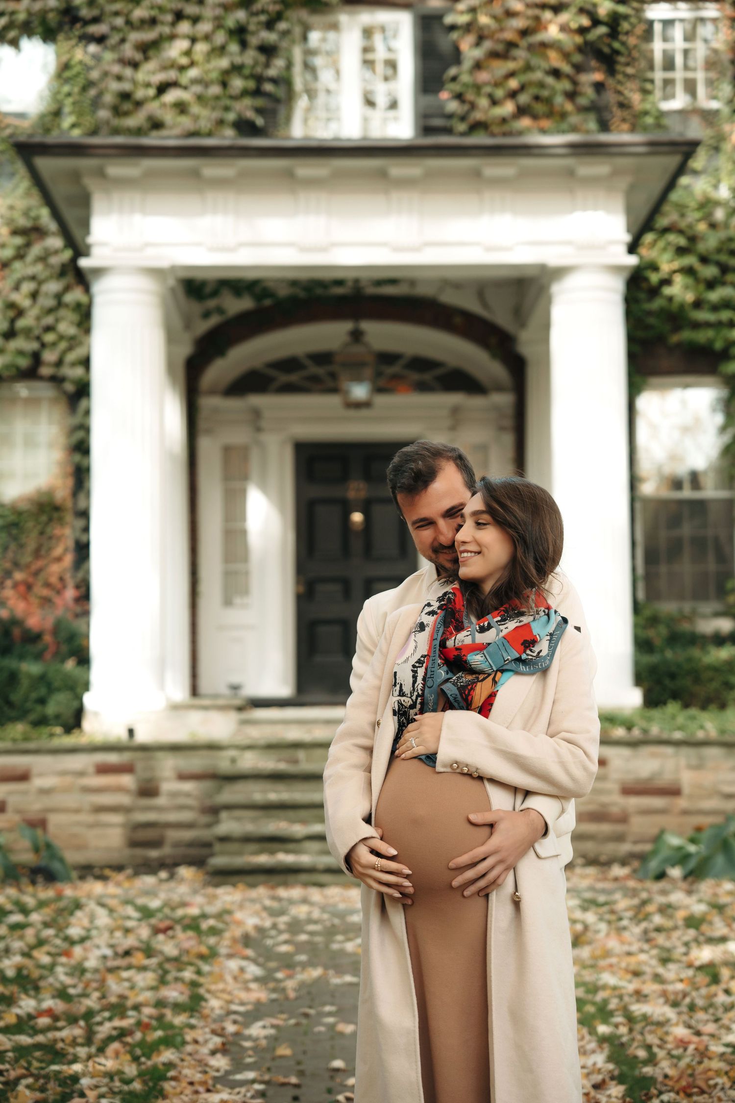 A couple shares an intimate autumn moment outside a historic white-columned entrance covered in fall foliage.