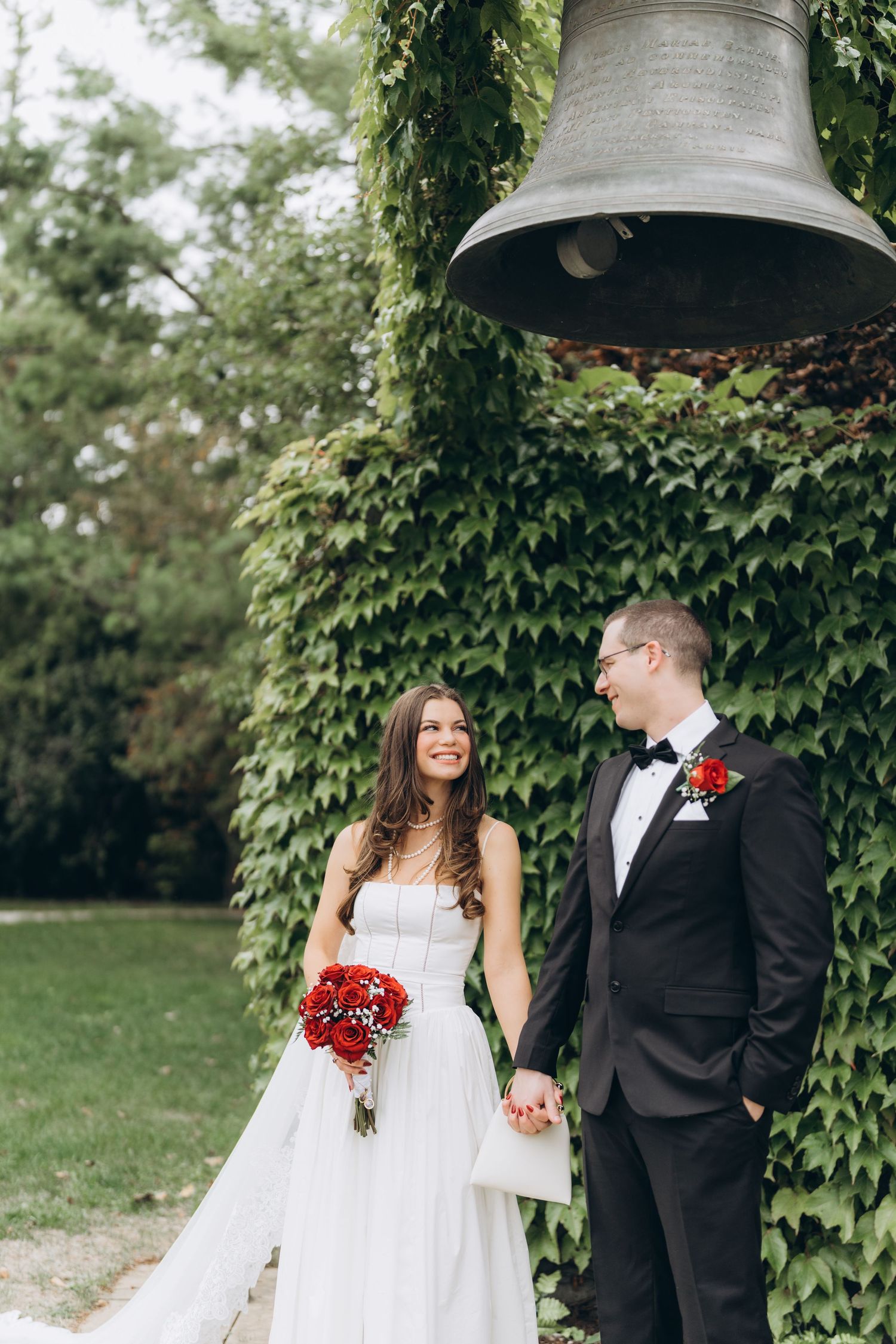 A couple poses together in their wedding attire against a lush green ivy wall backdrop during their outdoor celebration.