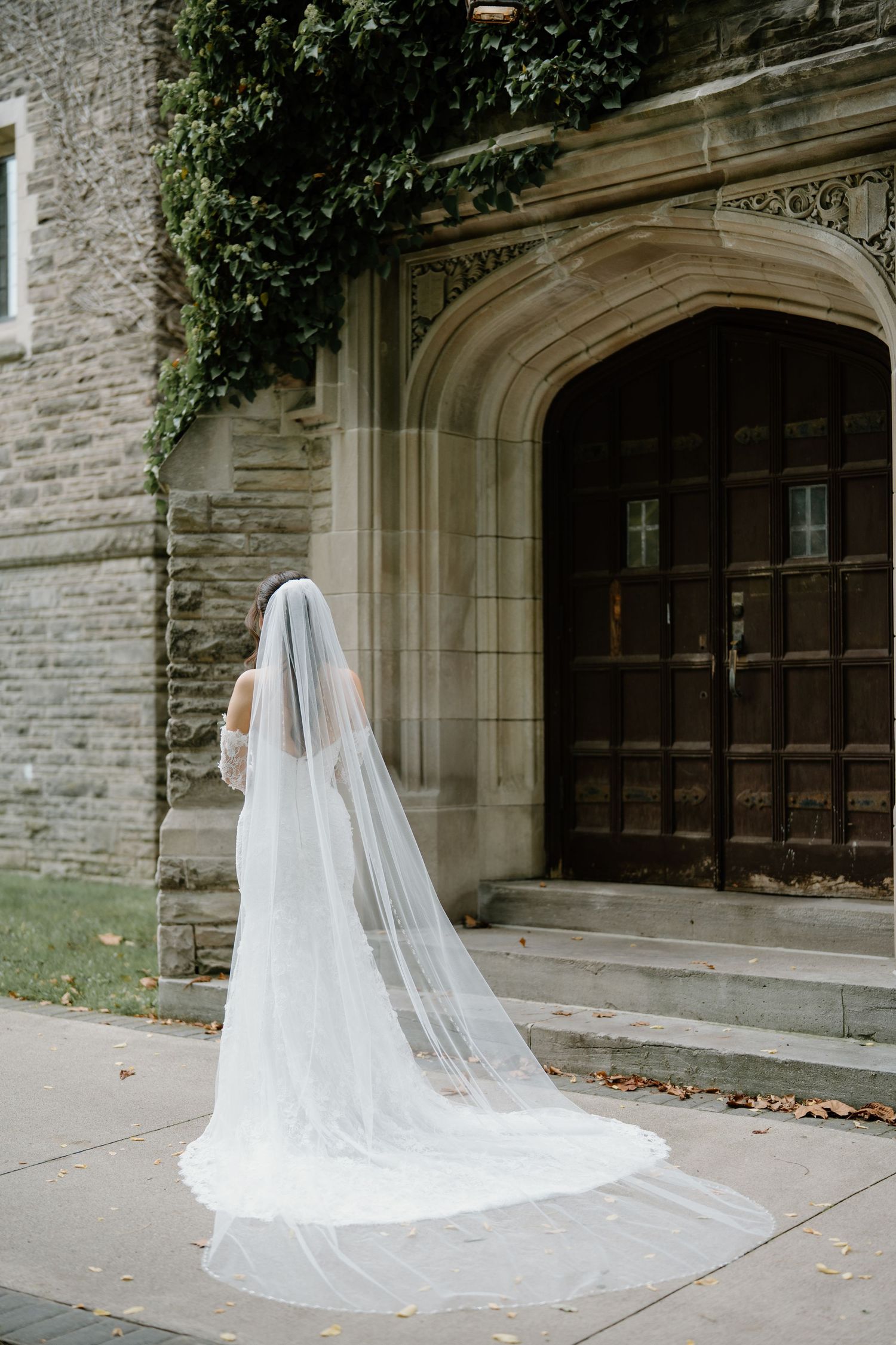 A cathedral length veil trails behind a bride as she stands before an ornate stone building entrance.