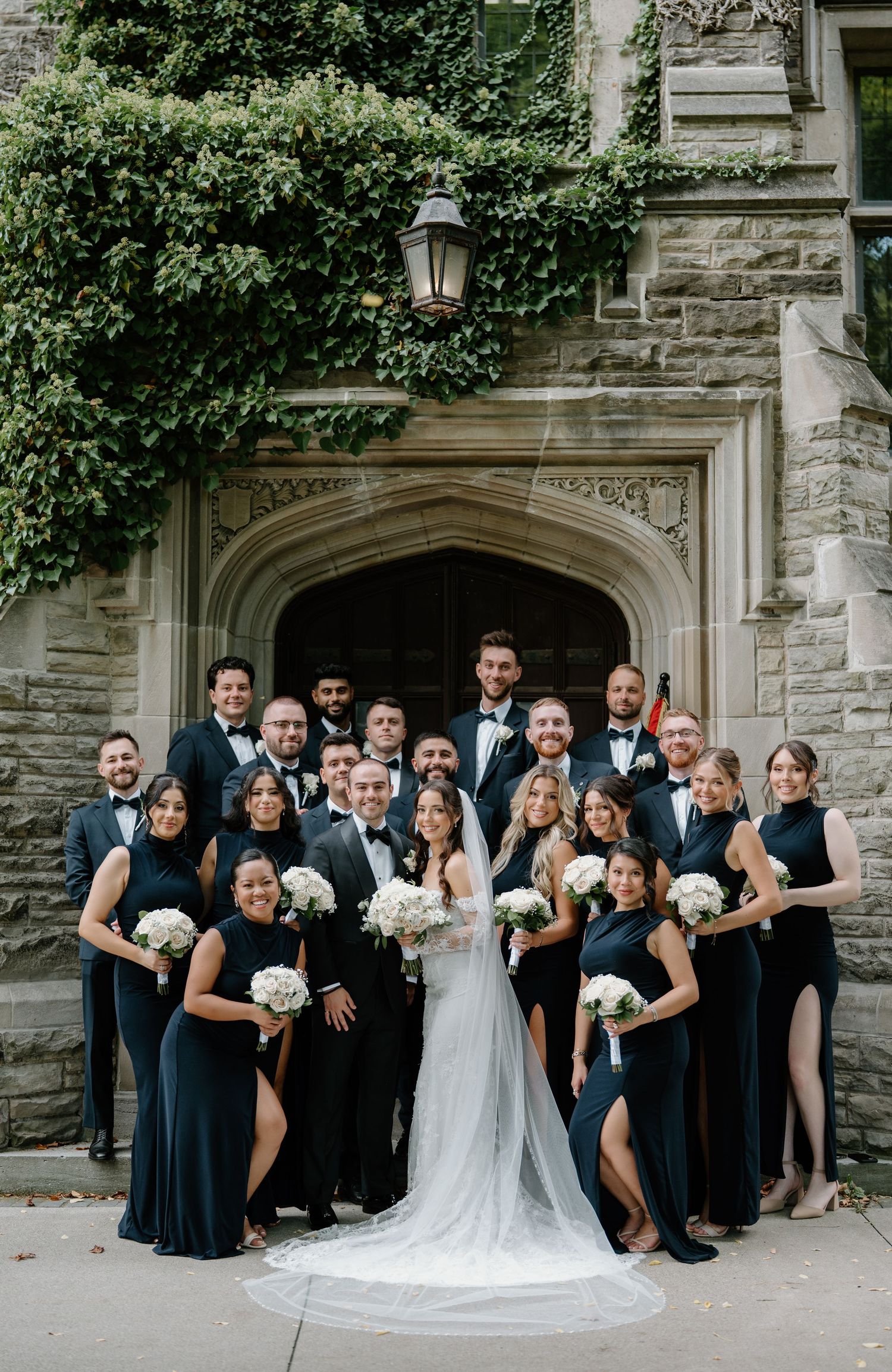 Wedding party in navy blue and white poses together in front of stone archway with greenery.