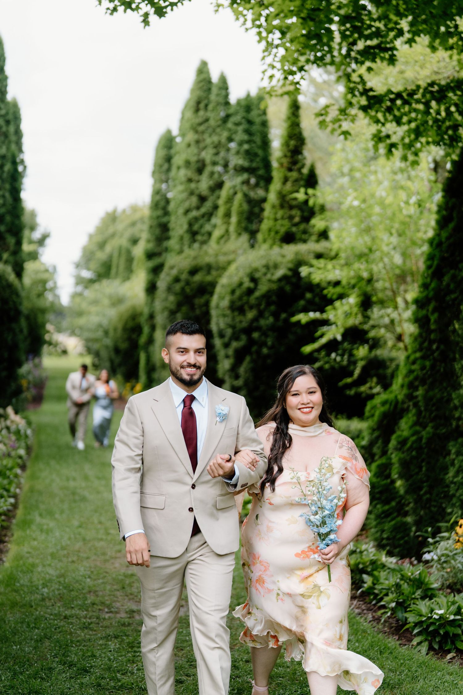 A couple walks along a lush garden path lined with tall cypress trees on a sunny day.