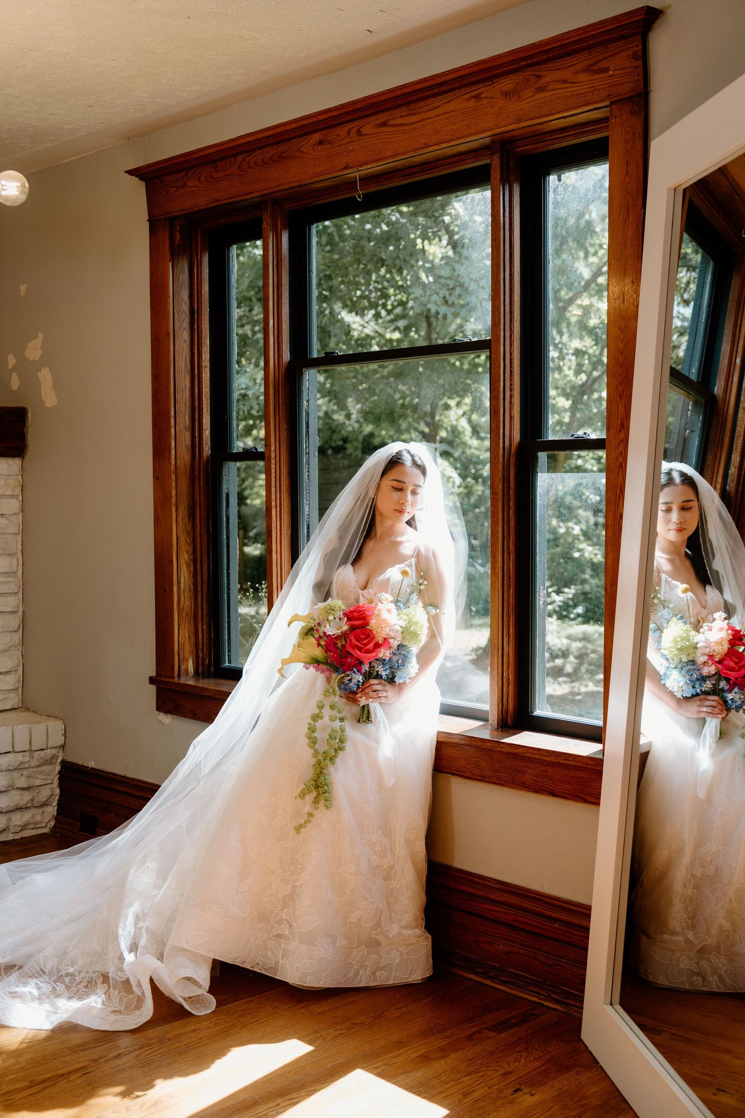 A bride in a white wedding dress poses by sunlit windows in a rustic room with wooden trim.