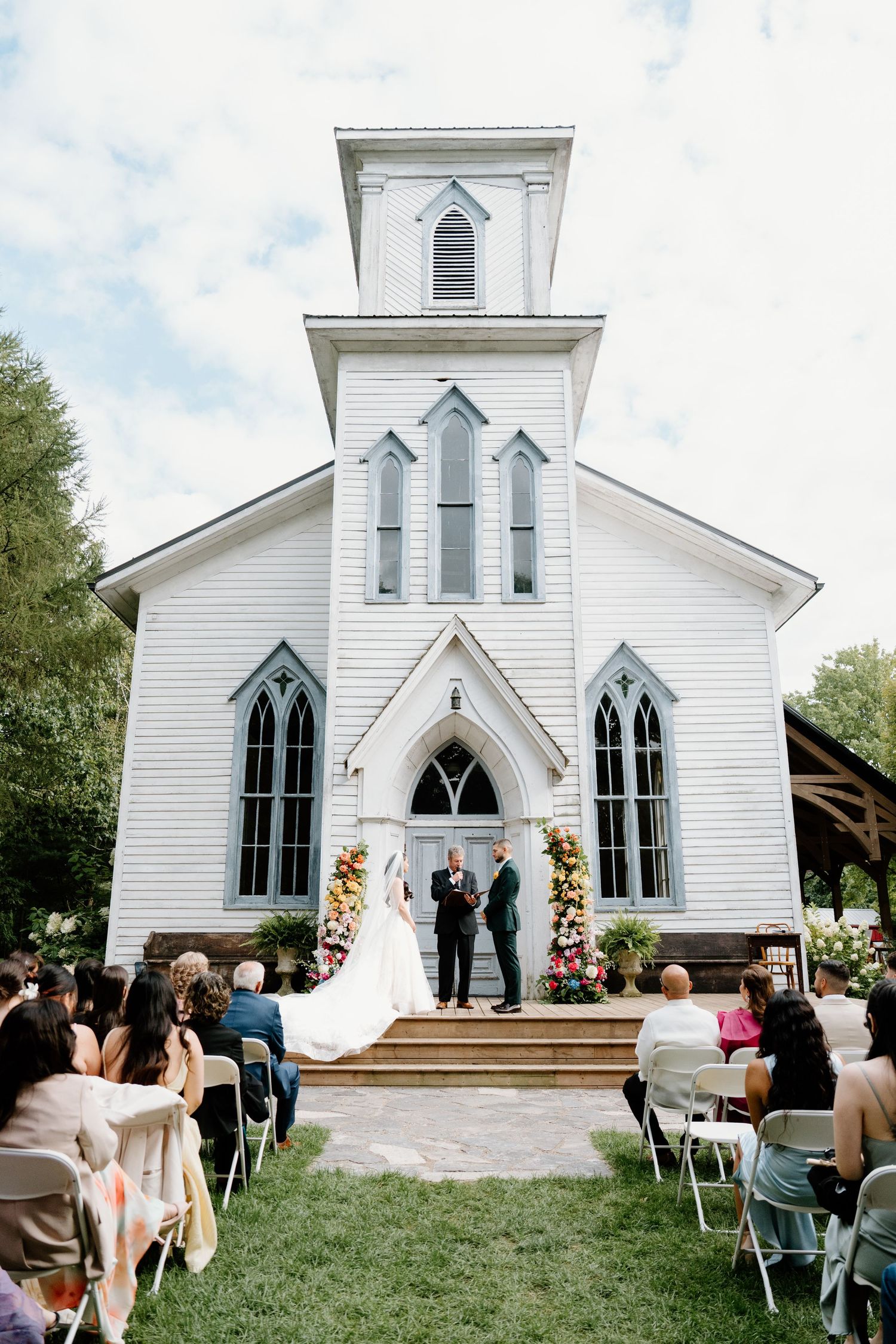A classic white country church with gothic windows hosts an outdoor wedding ceremony with guests seated on the lawn.