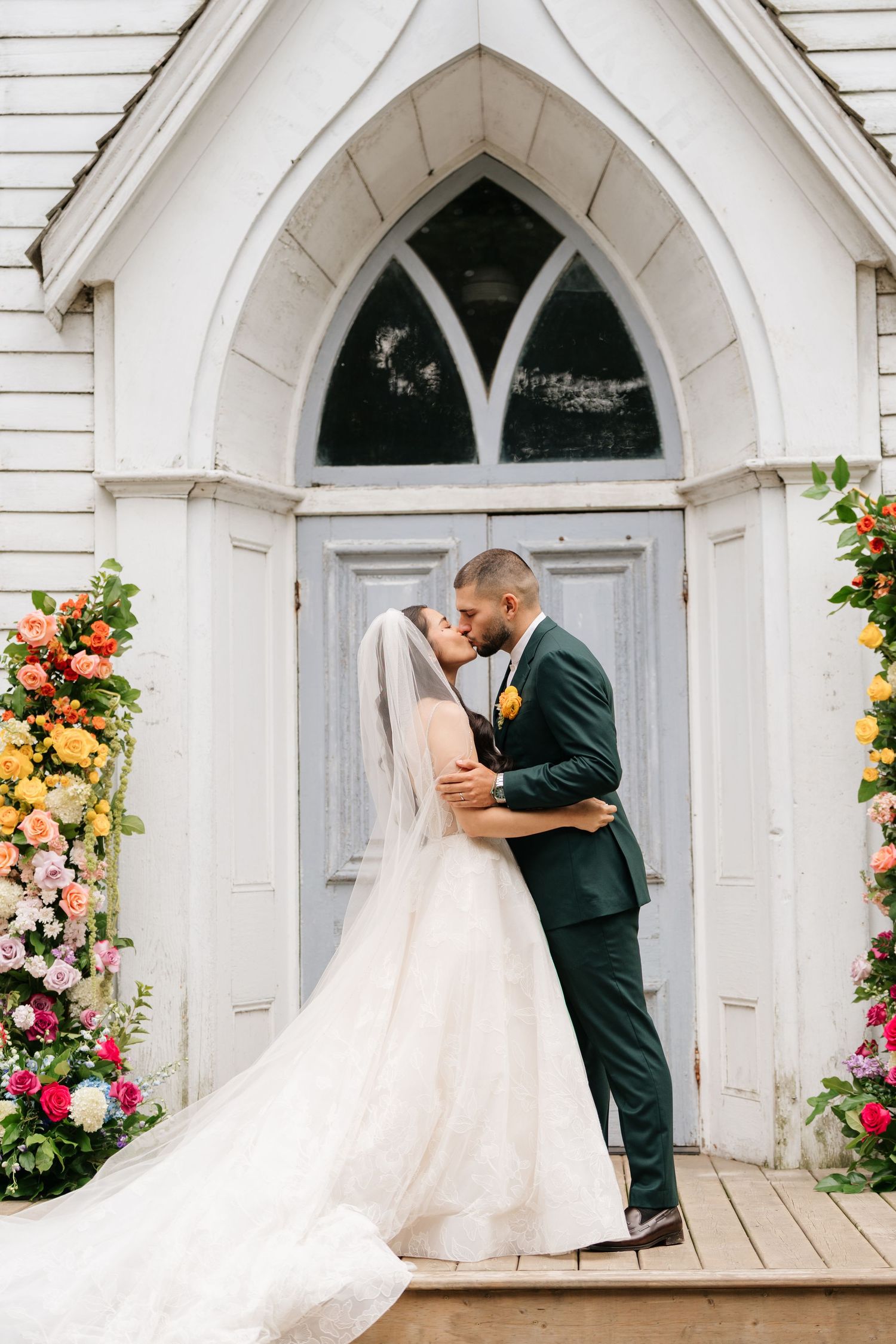 A couple shares a romantic moment in front of an elegant white church decorated with colorful flowers.