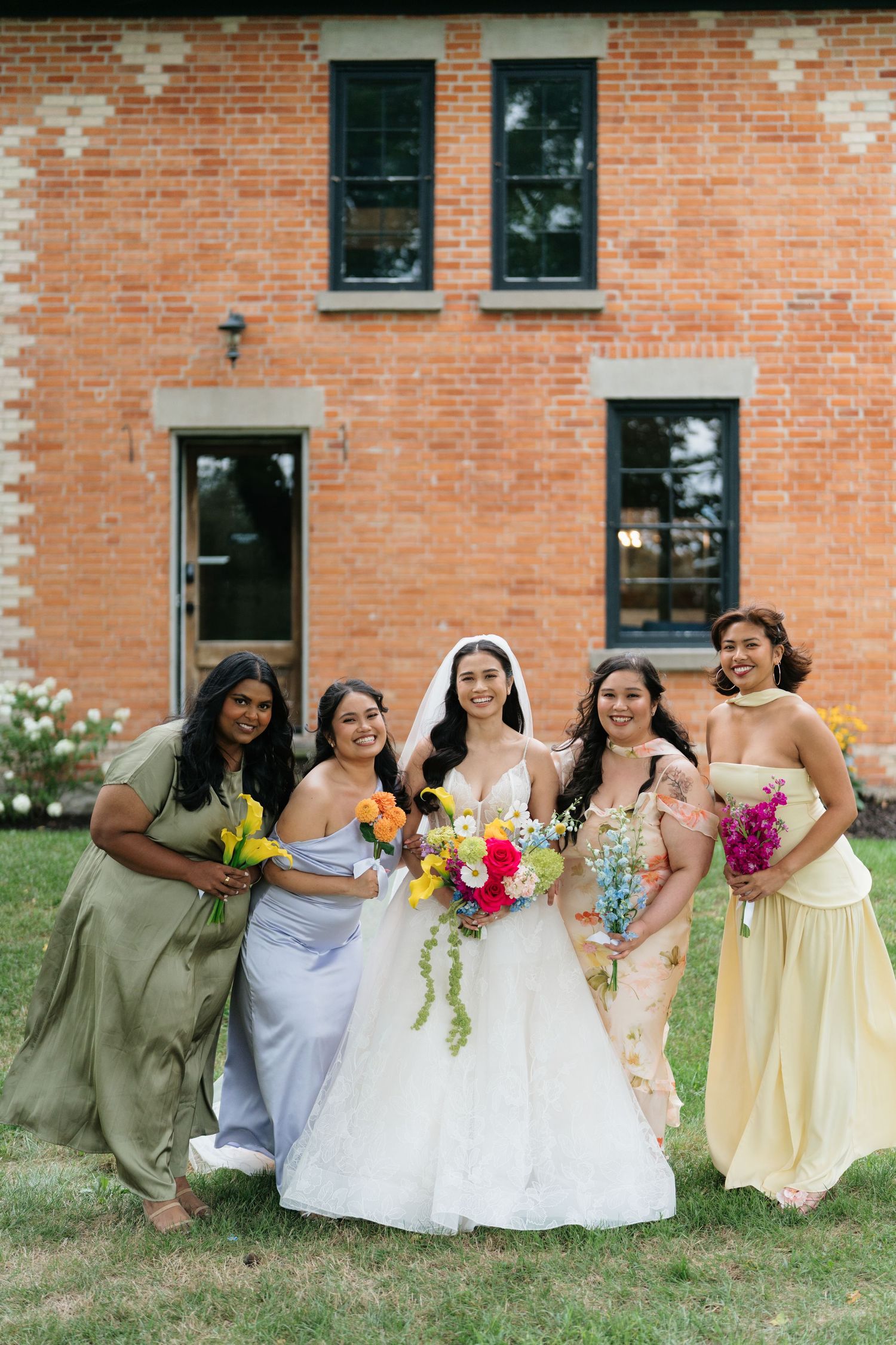 A wedding party in yellow dresses and light blue attire poses together on the grass by a brick building.
