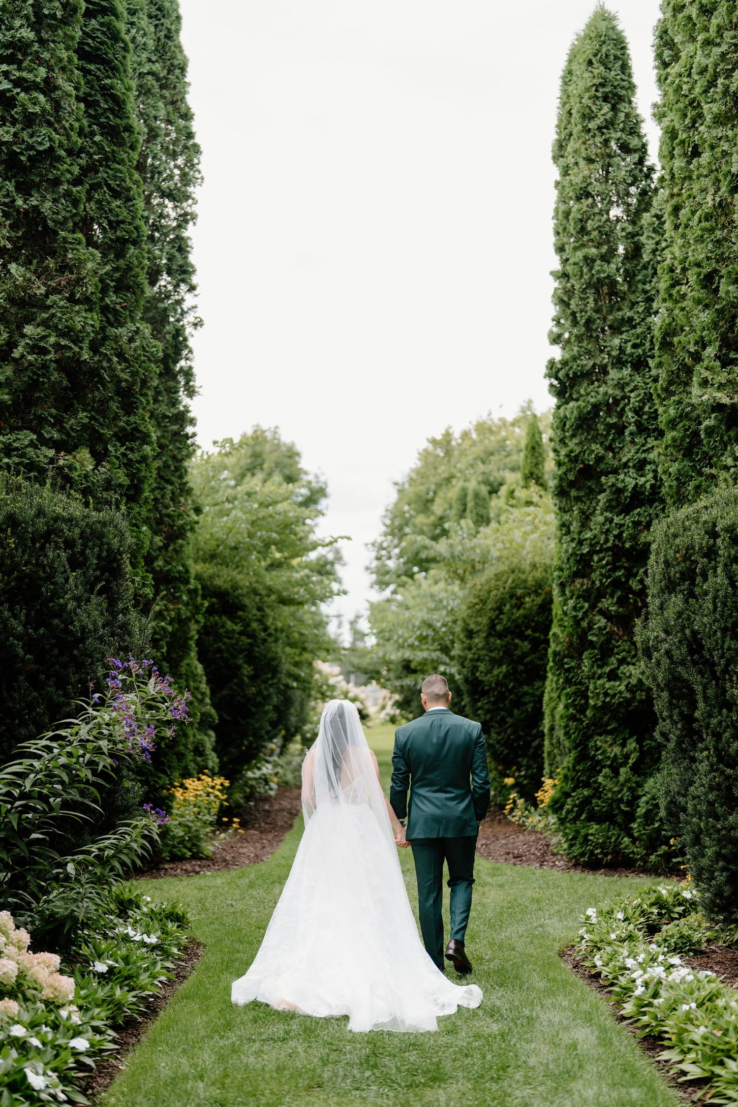 A couple walks down a garden path lined with tall cypress trees and colorful flowers on their wedding day.