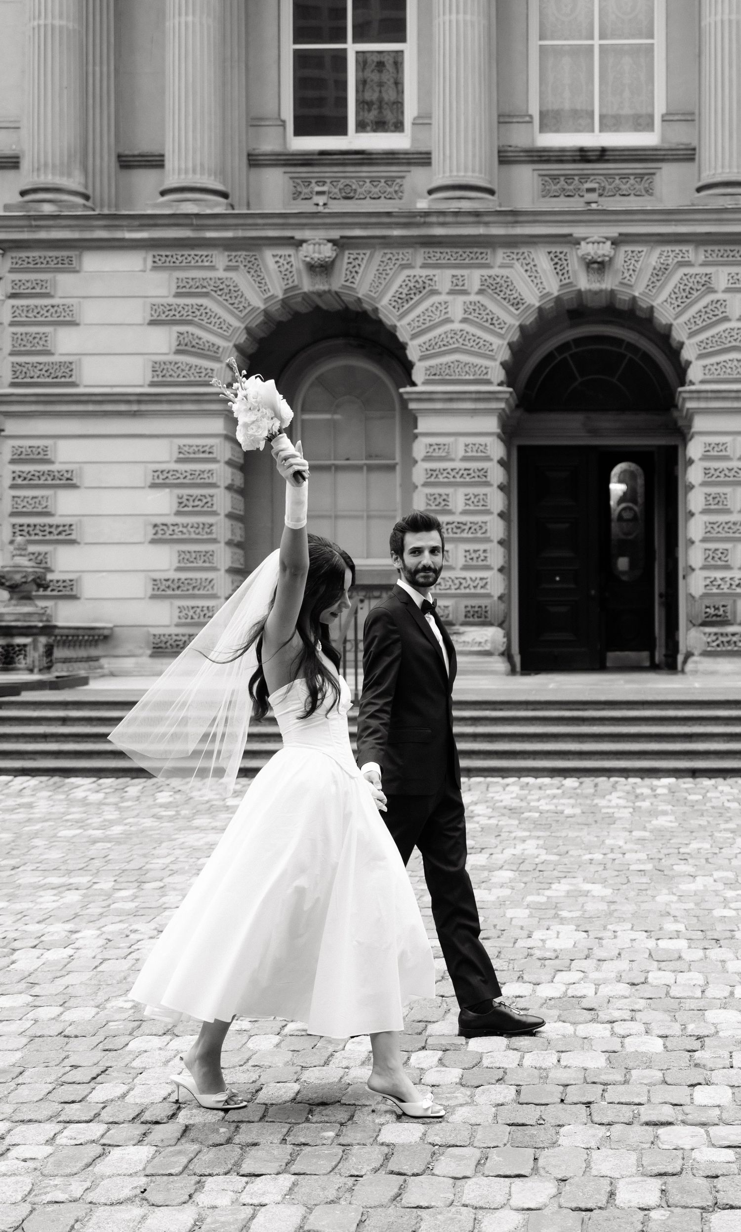 A romantic black and white wedding photo showing a couple in formal attire posing in front of a grand historic building.