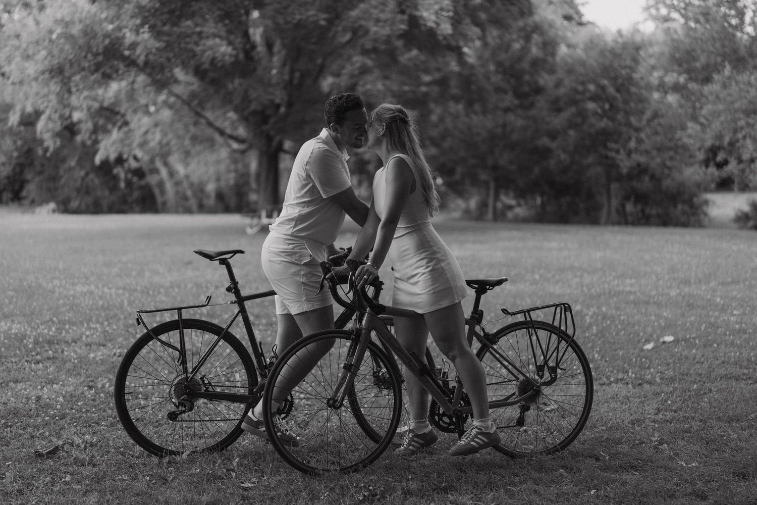 Two cyclists share a romantic moment with their bicycles in a park setting captured in black and white photography.