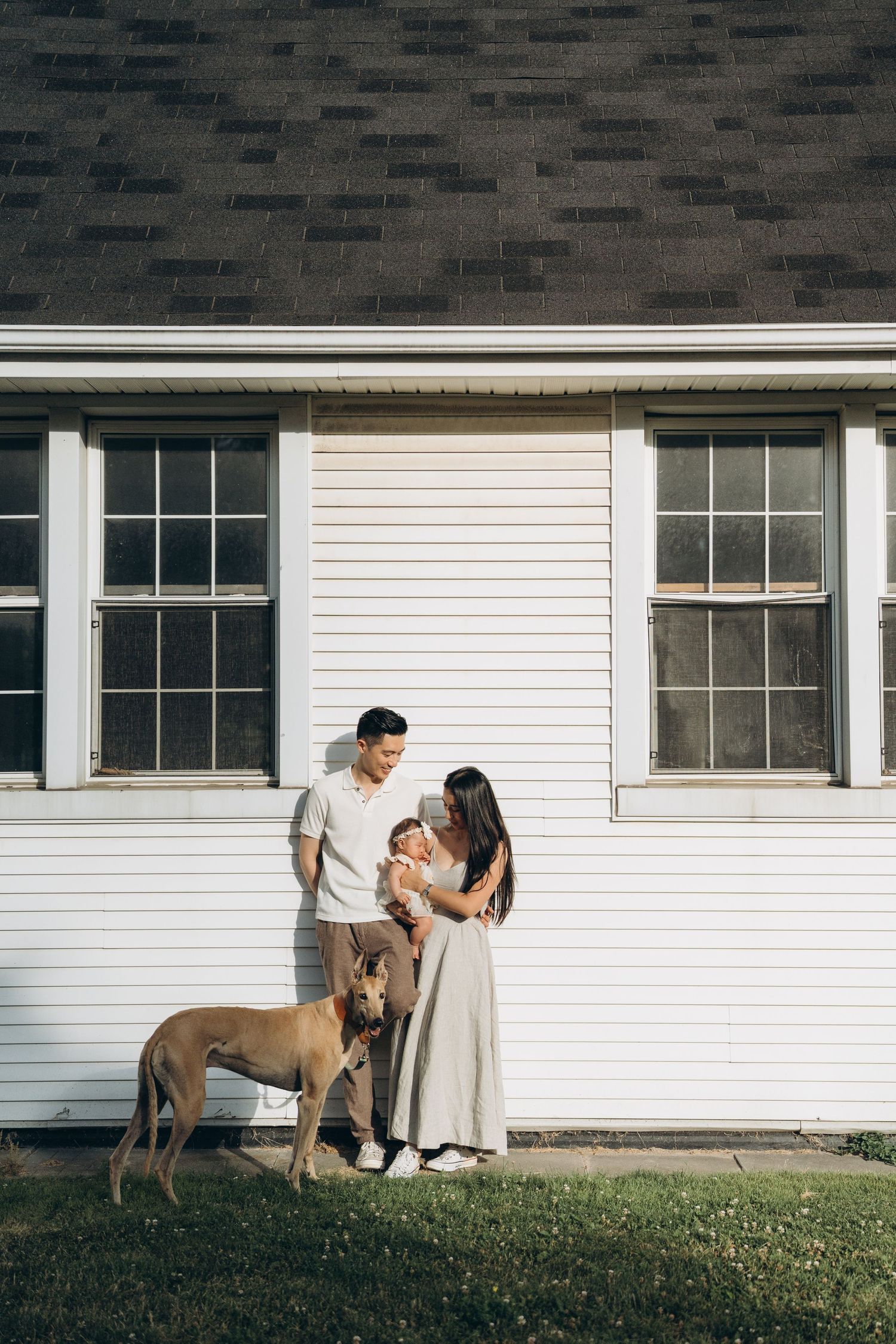 A couple stands with their brown dog in front of a white house with dark roof and windows.