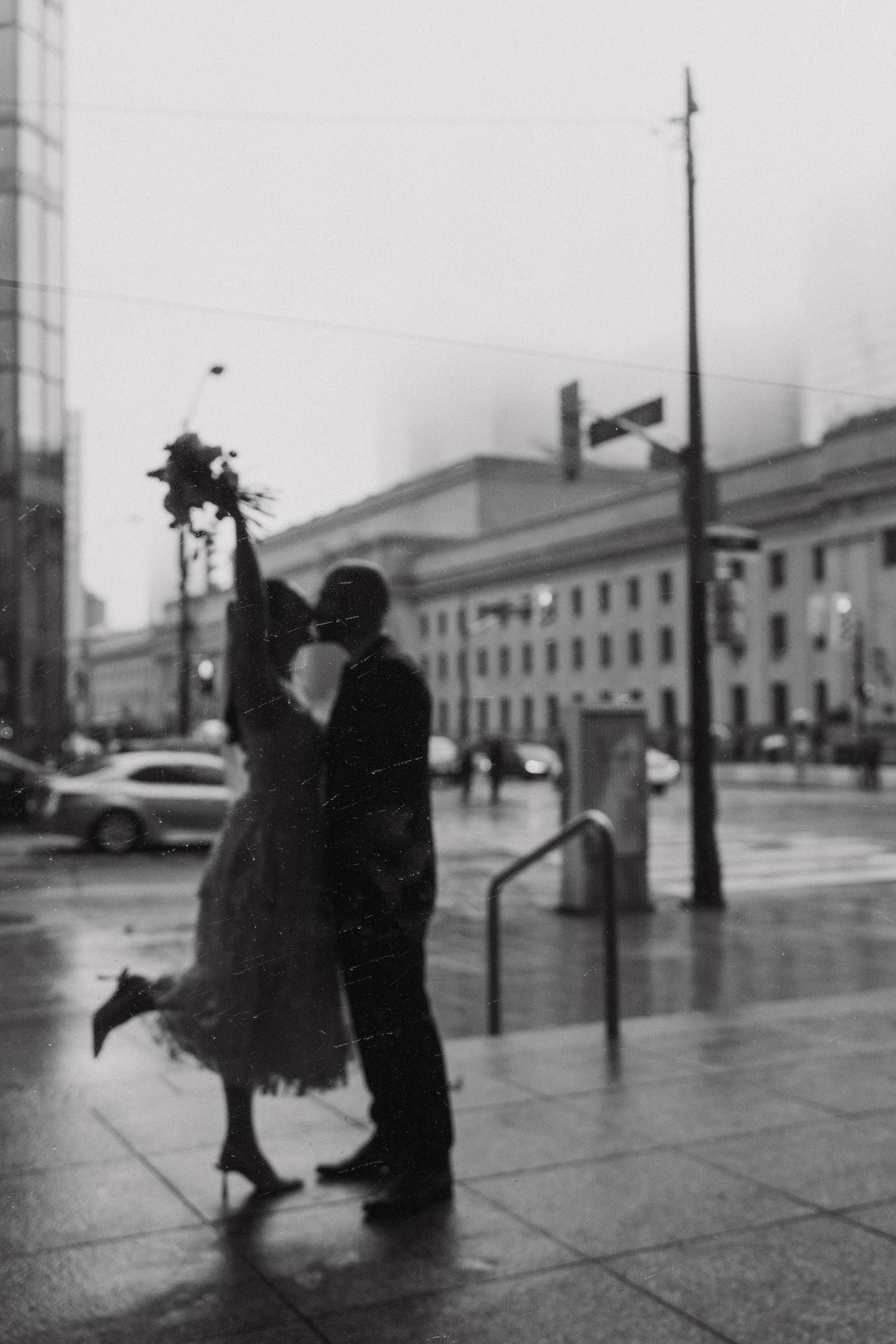 Black and white street scene showing silhouetted figures with umbrellas walking on a rainy city sidewalk.
