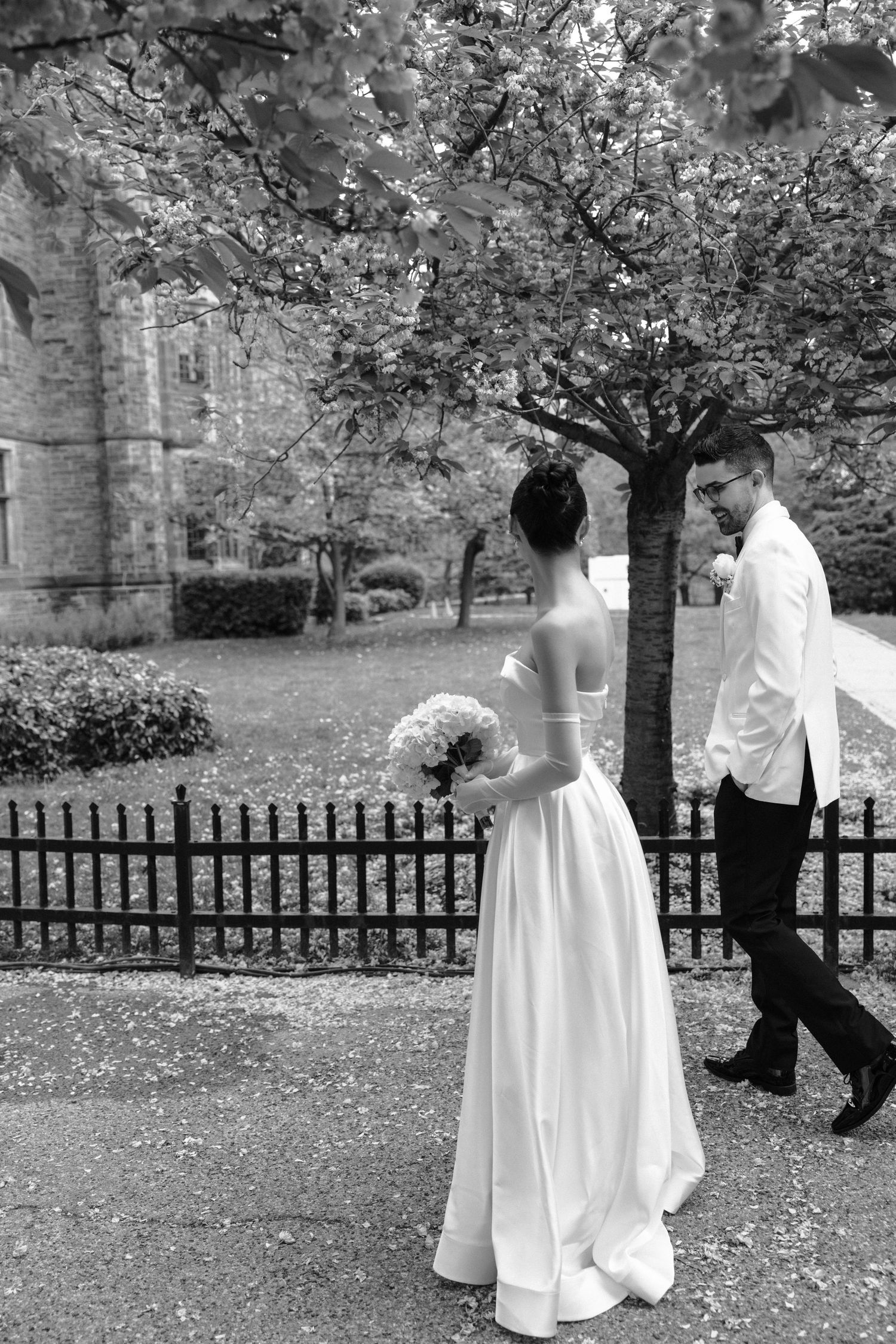 A romantic black and white wedding photo showing a couple in formal attire standing near an ornate iron fence in a garden.