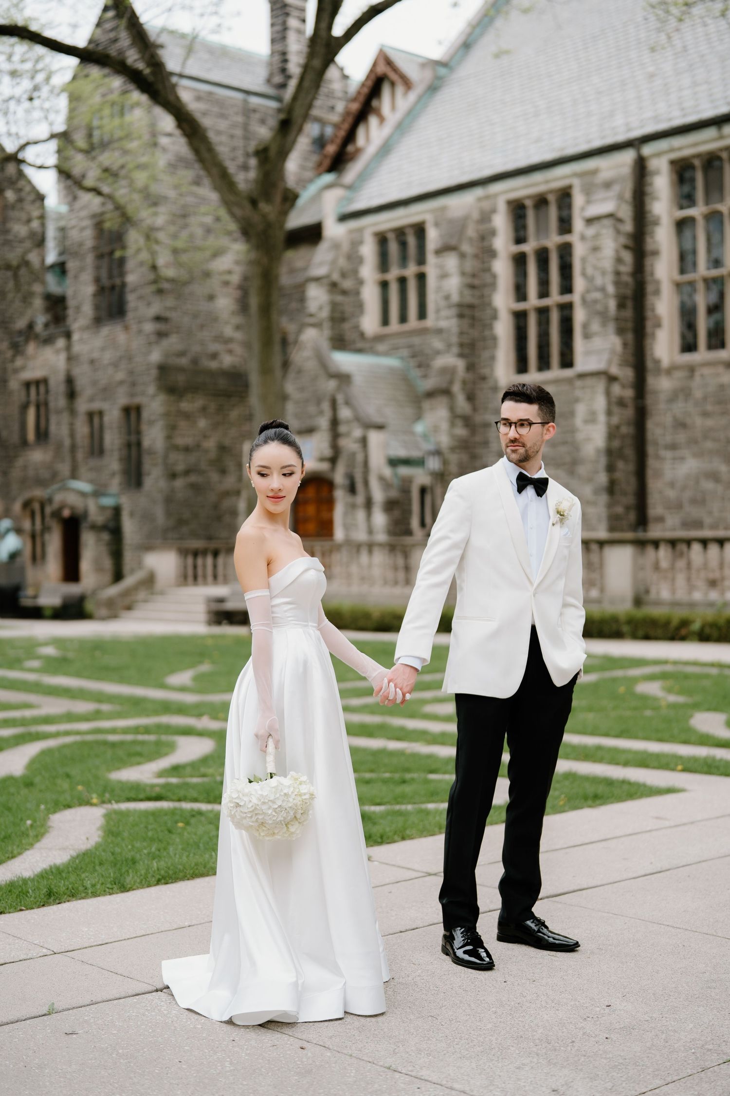 Elegant couple in formal wedding attire posing outside a Gothic stone building on a spring day.