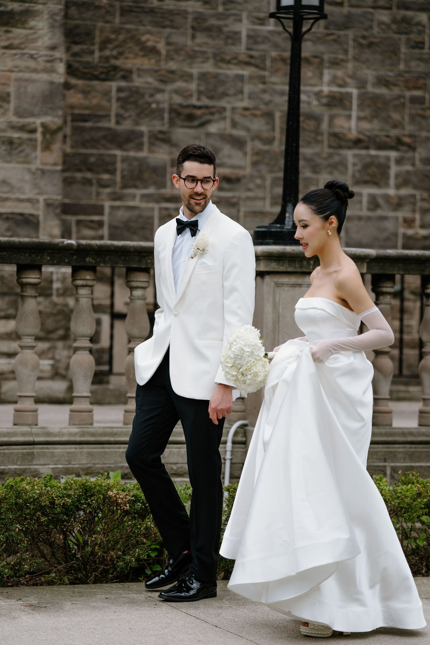 Elegant wedding couple poses against stone wall backdrop in classic black and white attire.