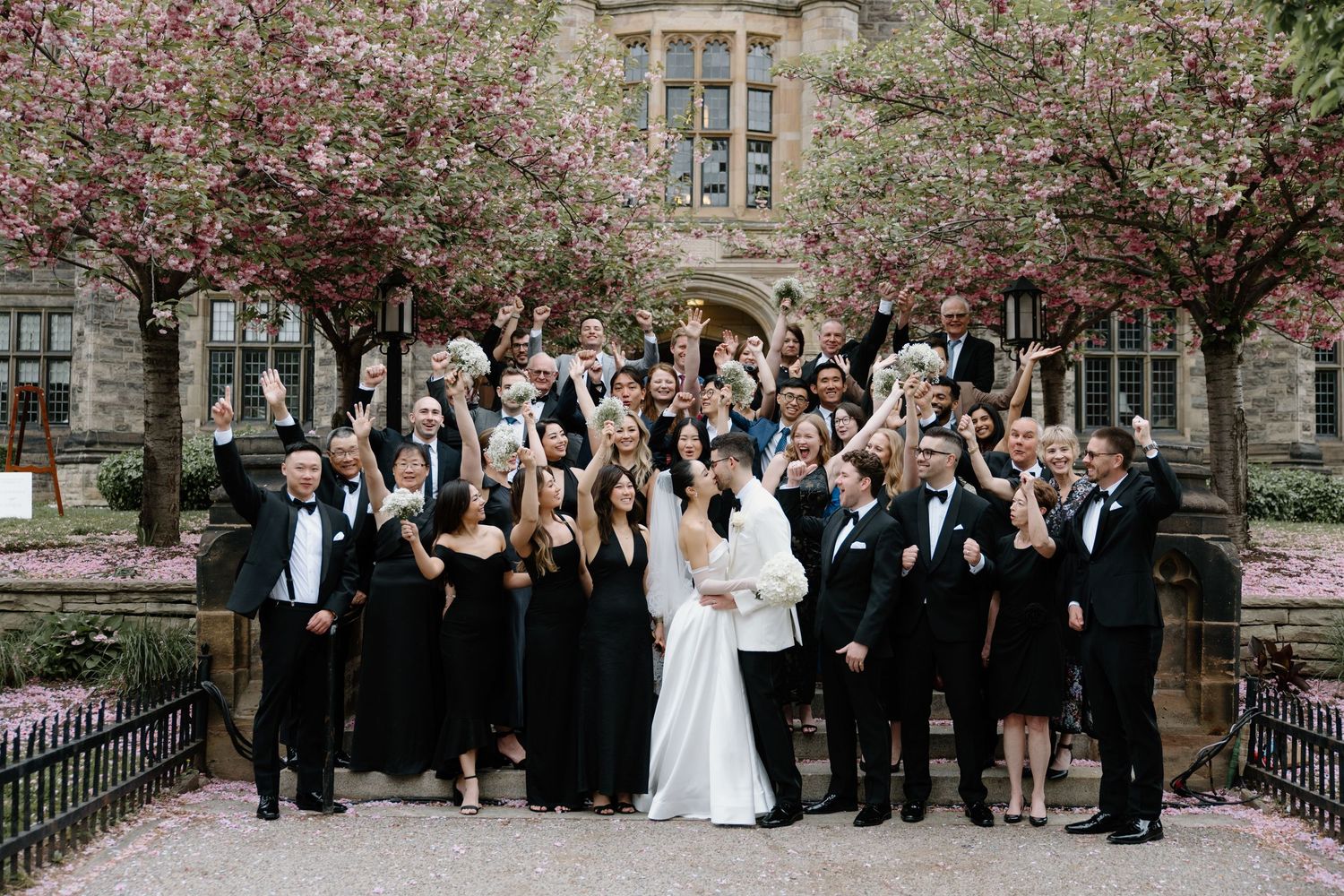 A wedding party poses for formal photos in a garden setting with blooming cherry blossoms and stone architecture.
