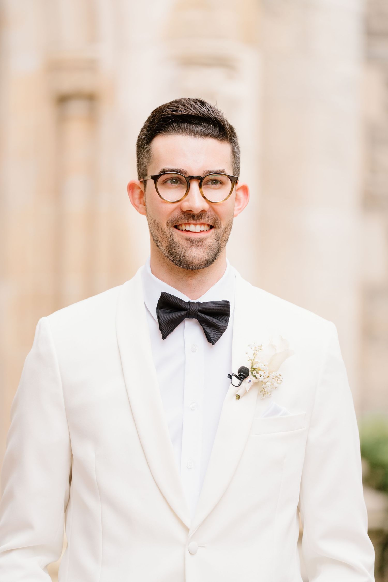 A person wearing a crisp white tuxedo jacket, bow tie and black rimmed glasses smiles warmly at the camera.