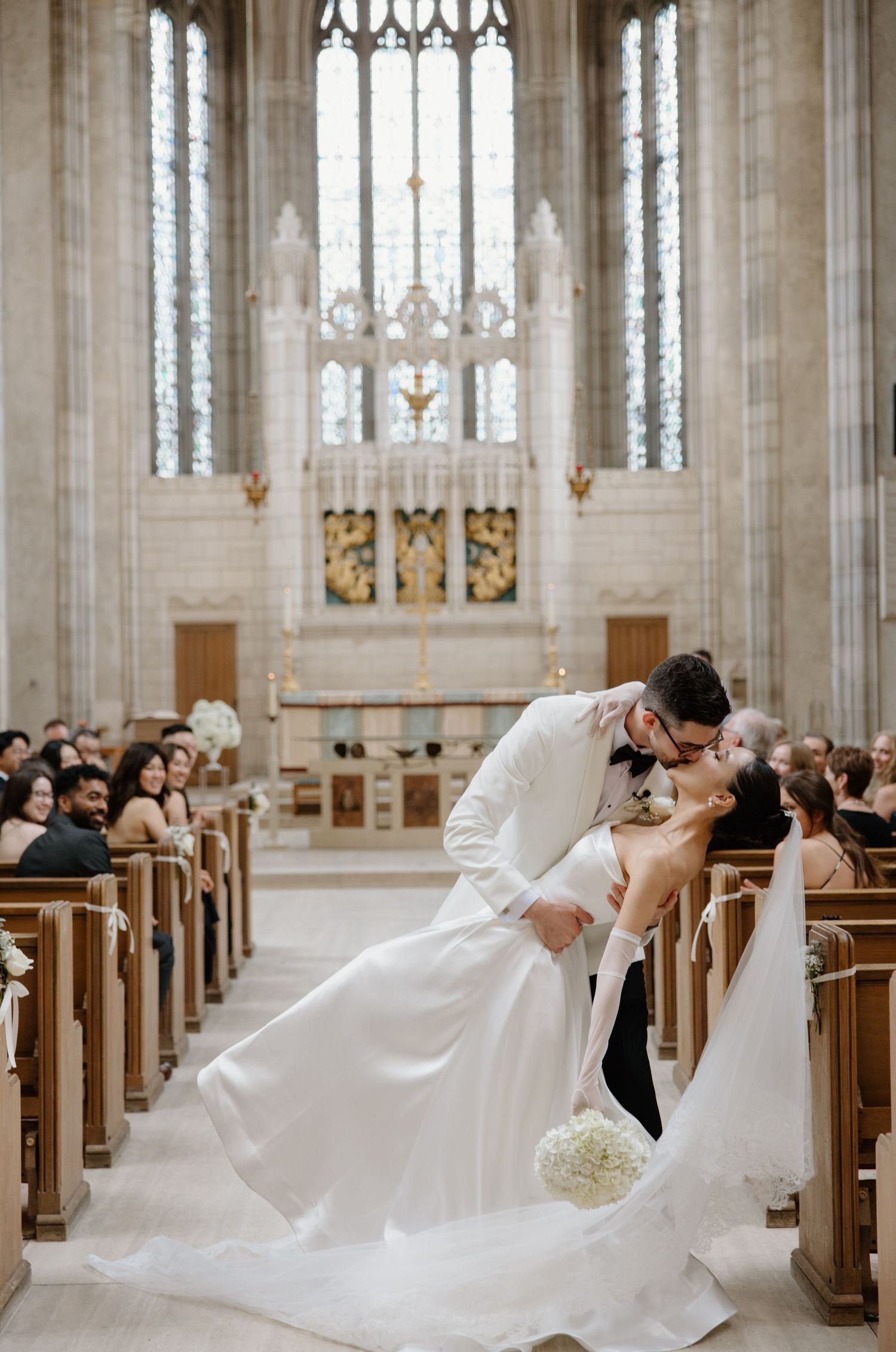 A romantic wedding moment captured in a grand cathedral with gothic architecture, stained glass windows and wooden pews filled with guests.