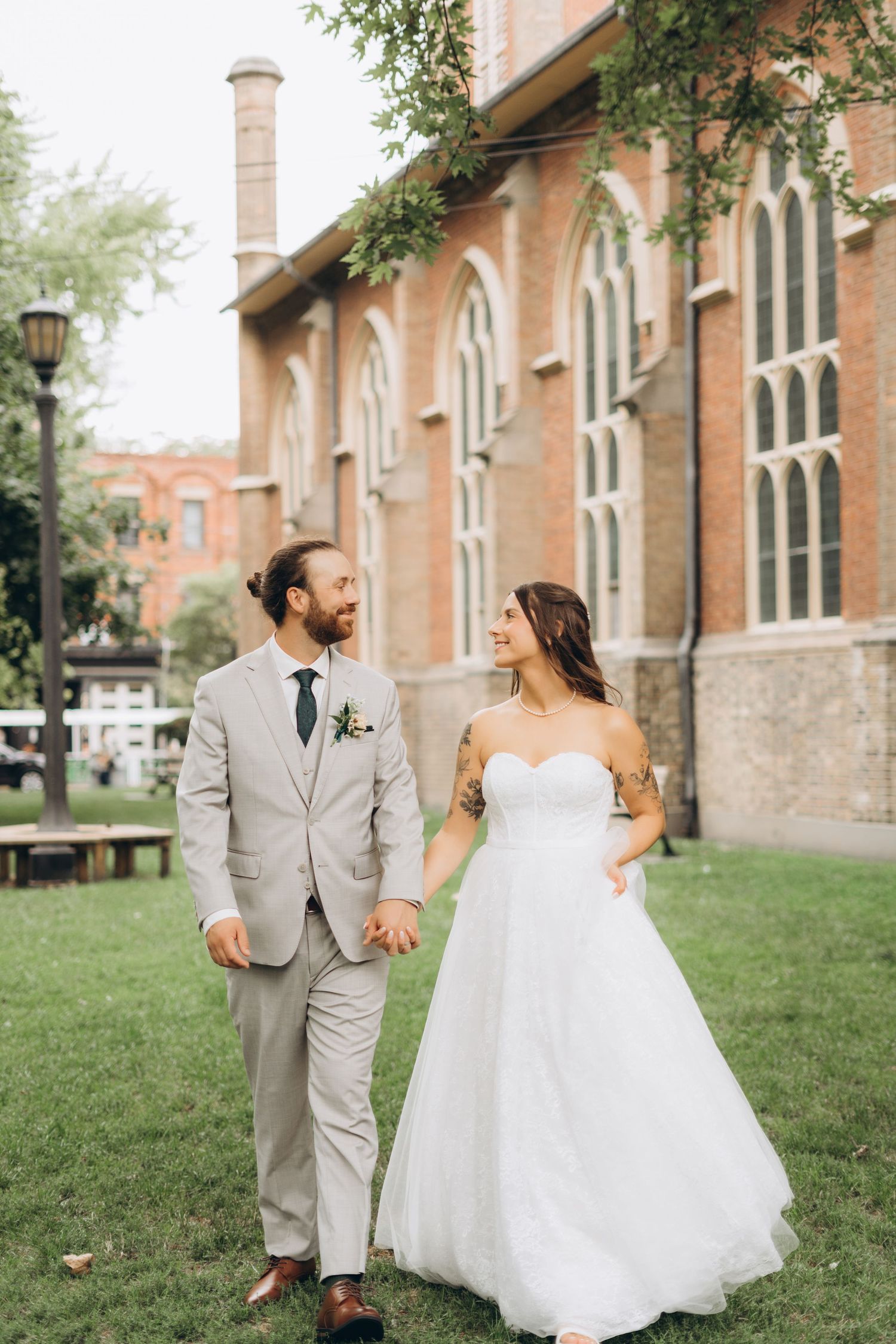 A couple in wedding attire walks together on grass outside a historic brick church building with arched windows.