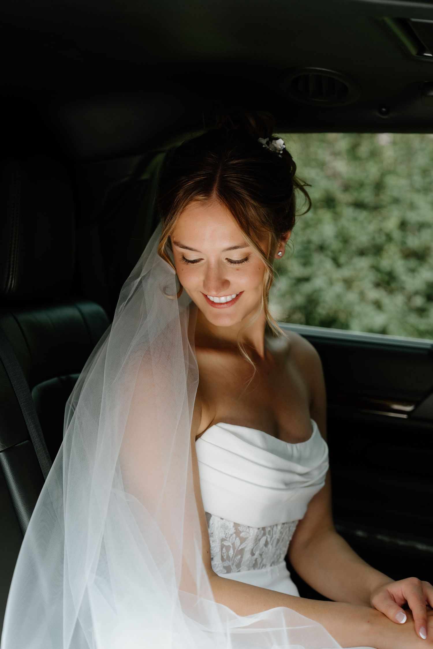 A bride in a white off-shoulder wedding dress sits in a car with natural light illuminating her veil and smile.