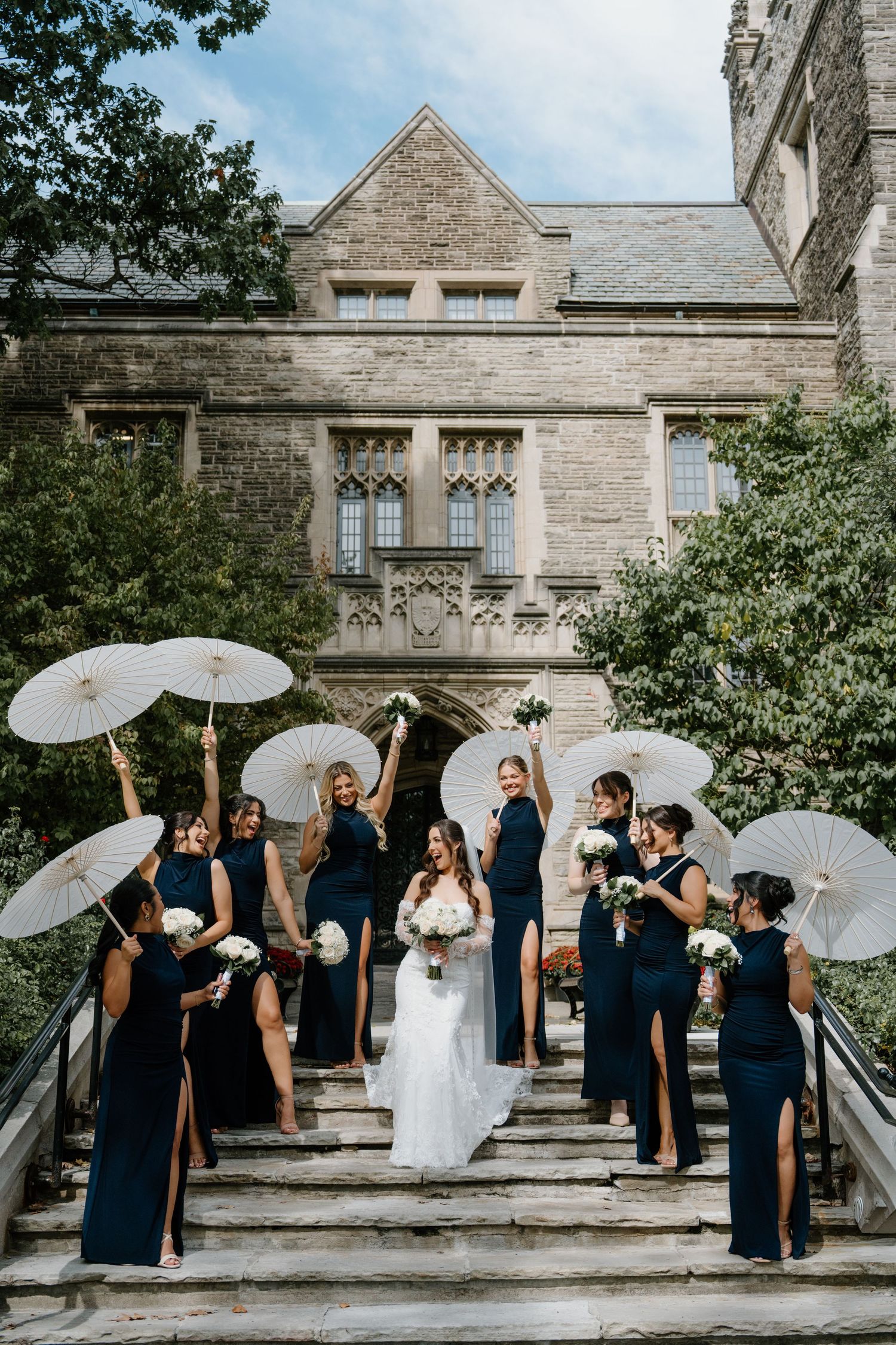 Wedding party poses with white parasols against stone building backdrop in playful sequence of candid moments.