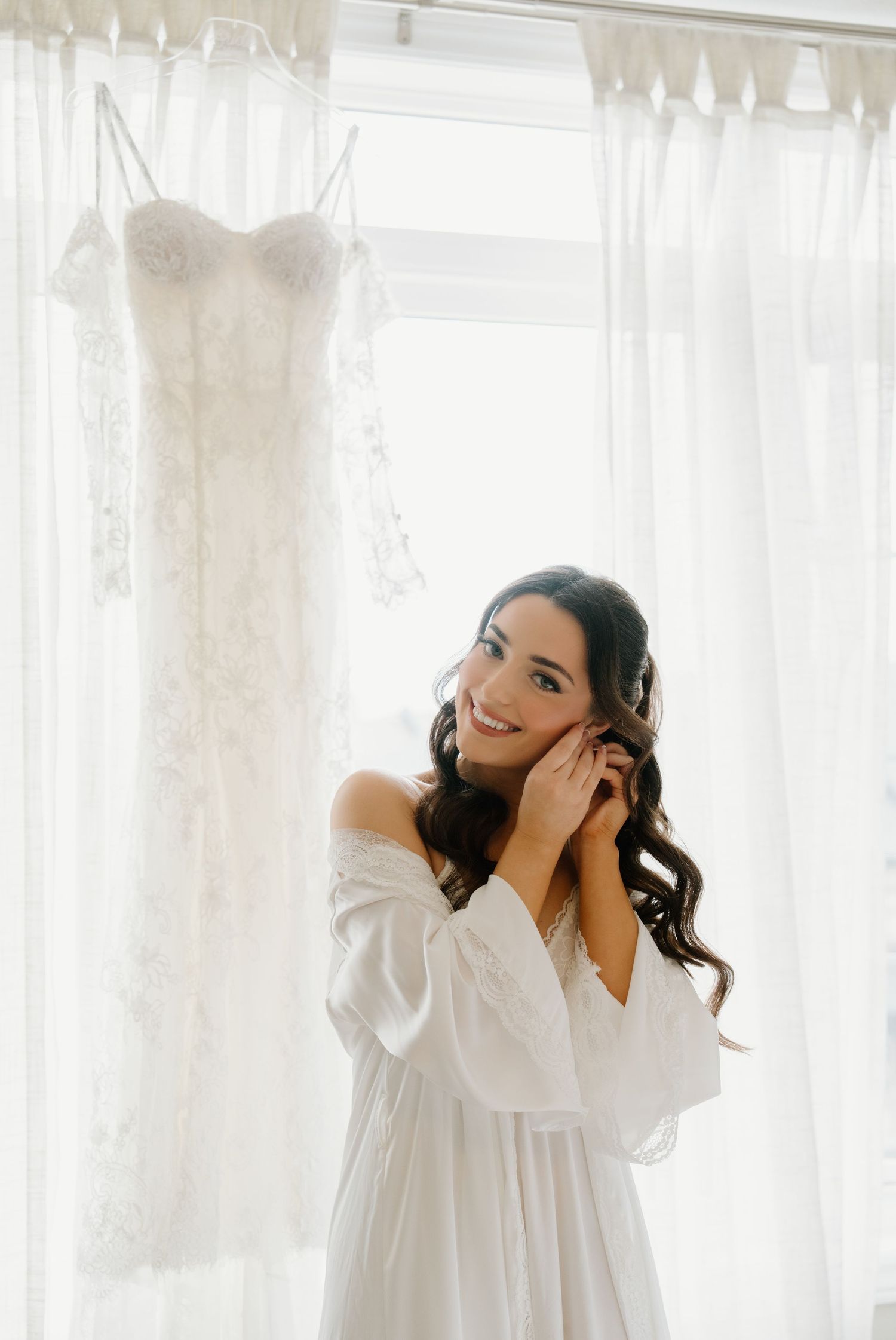 Woman in white dress poses gracefully against sheer curtains in a bright interior setting.