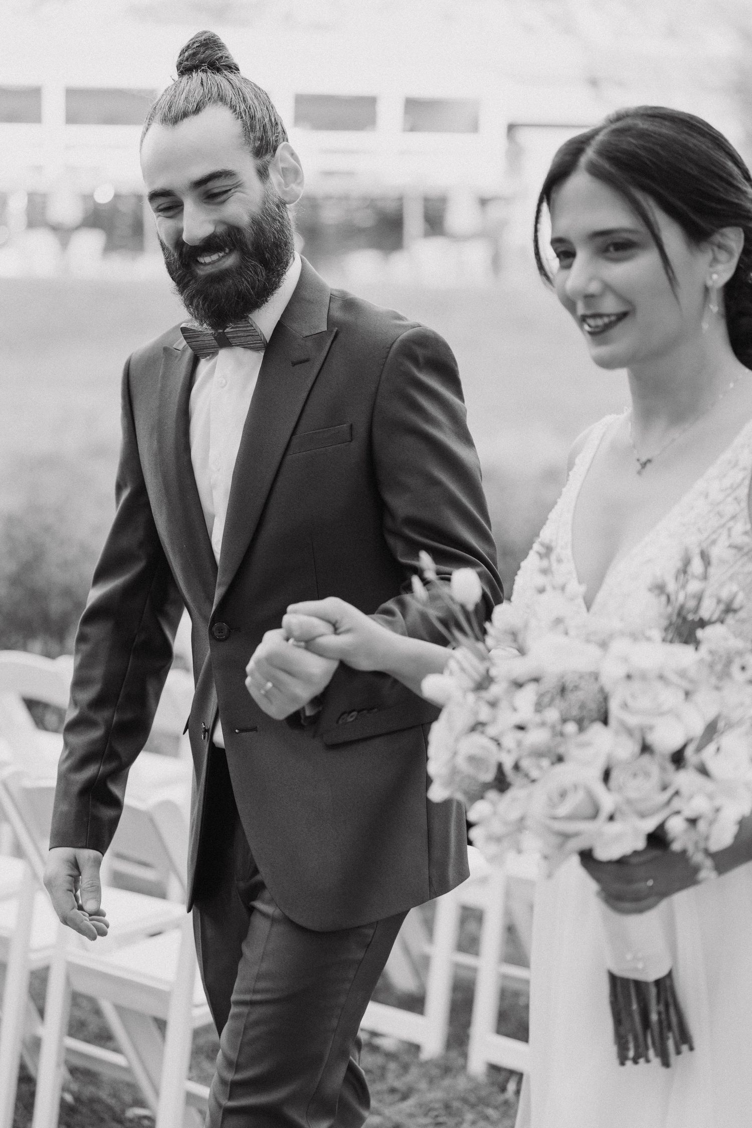 Black and white candid moment of newlyweds walking together, with bride holding floral bouquet.