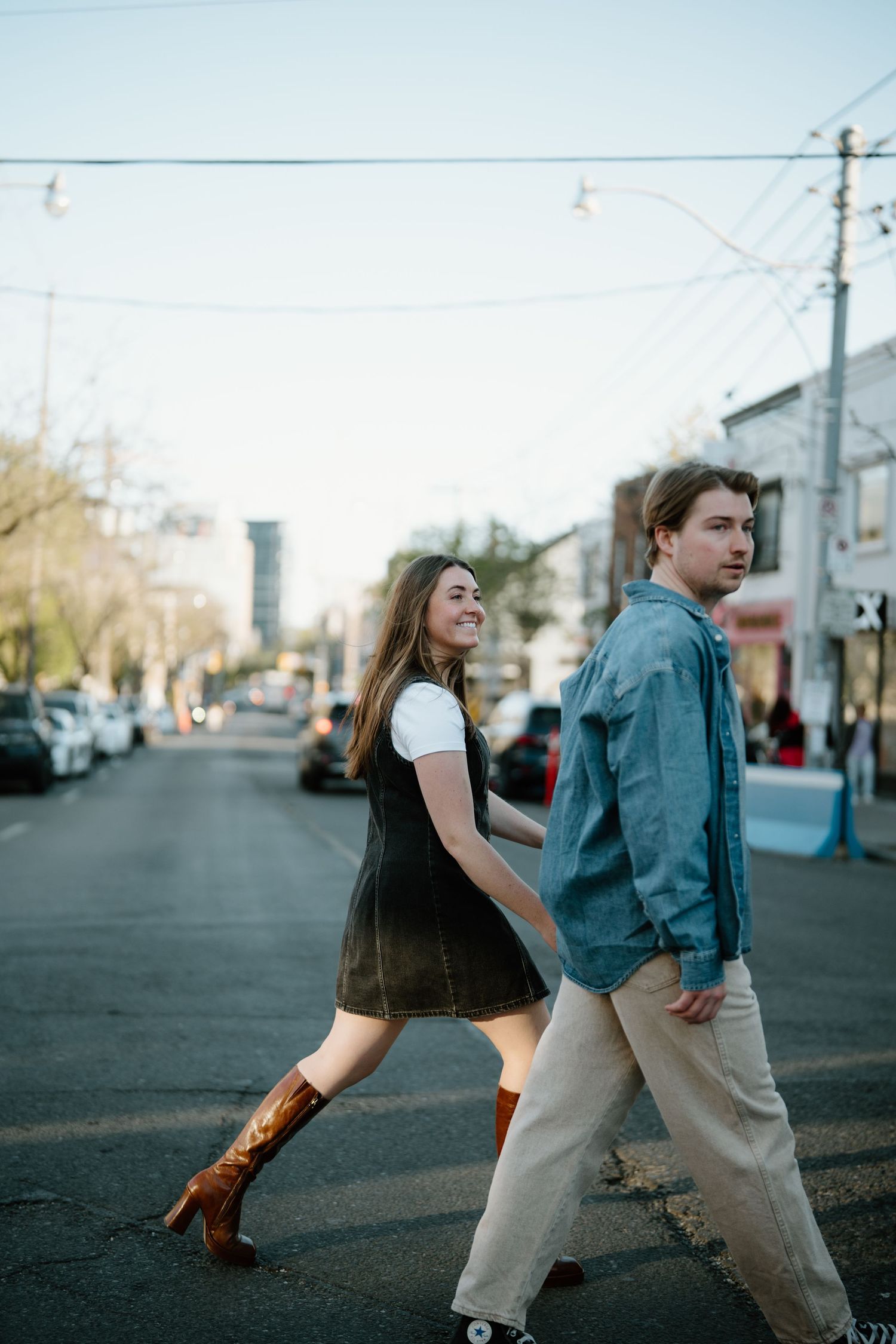 A young couple walks together on an urban street during golden hour, with her wearing brown boots and him in casual attire.