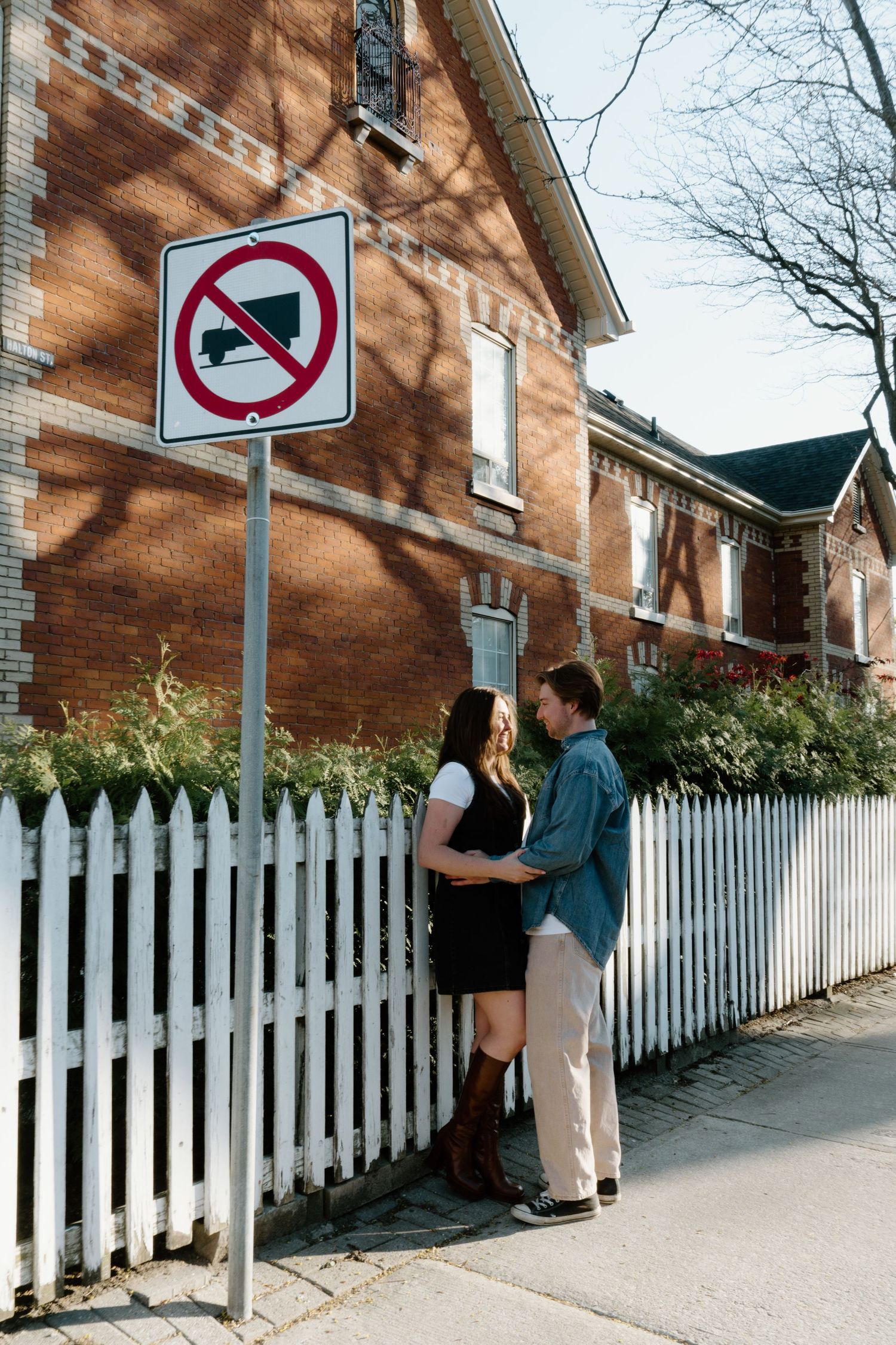 Two people share an intimate moment beside a white picket fence and brick house with a no honking traffic sign above.