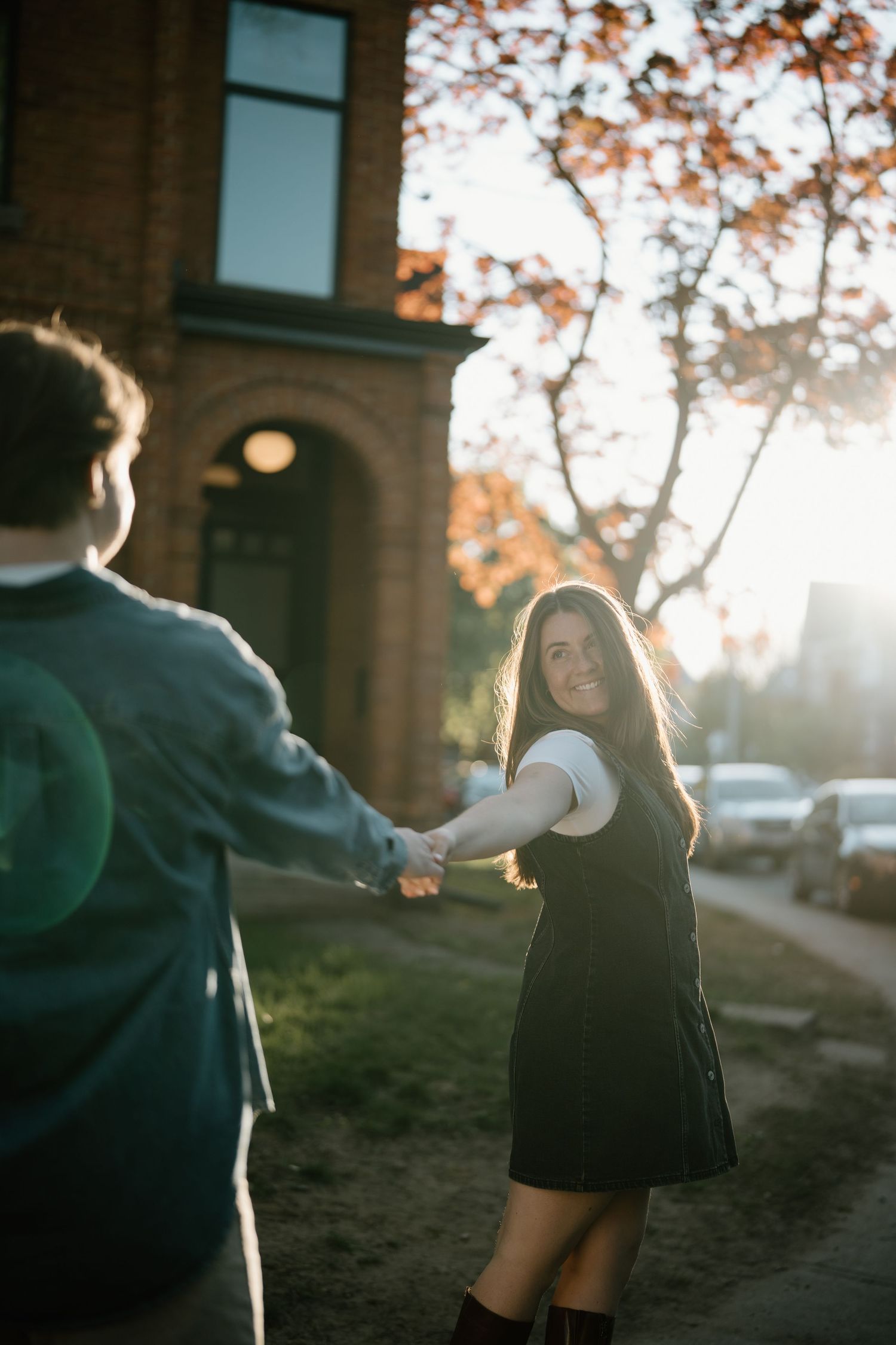 Two people hold hands and dance together on a sunlit autumn sidewalk with dramatic backlit lighting at golden hour.