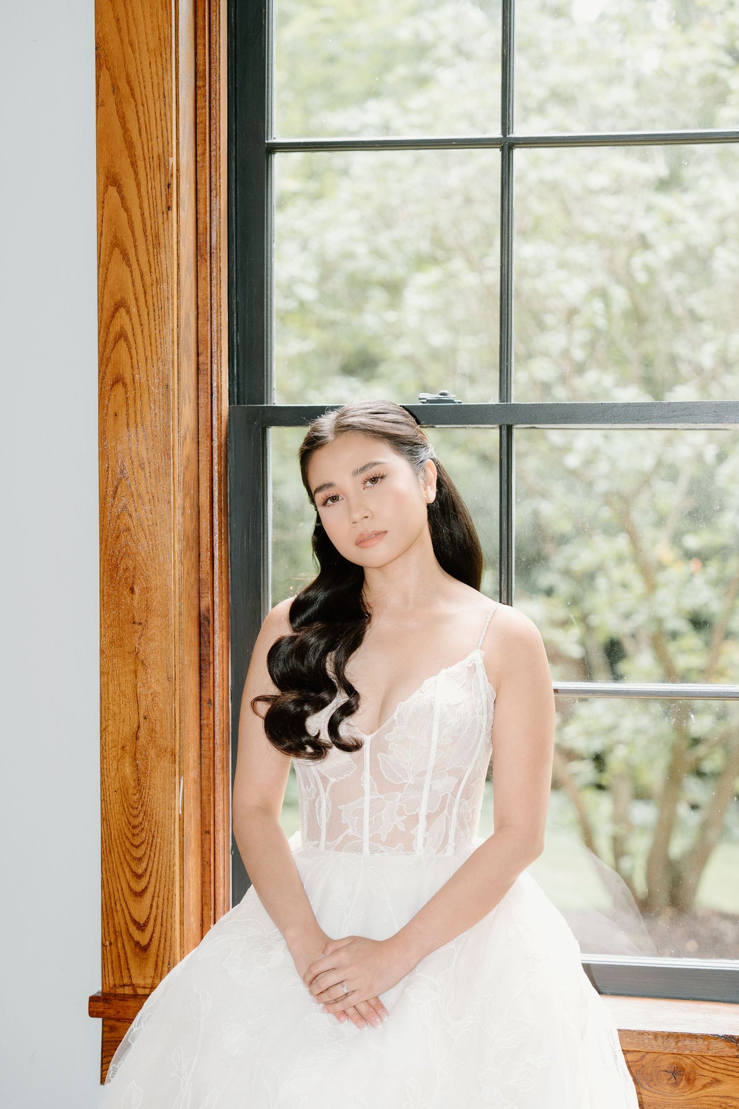 Bride in white gown poses elegantly by window with wooden trim in natural light setting.