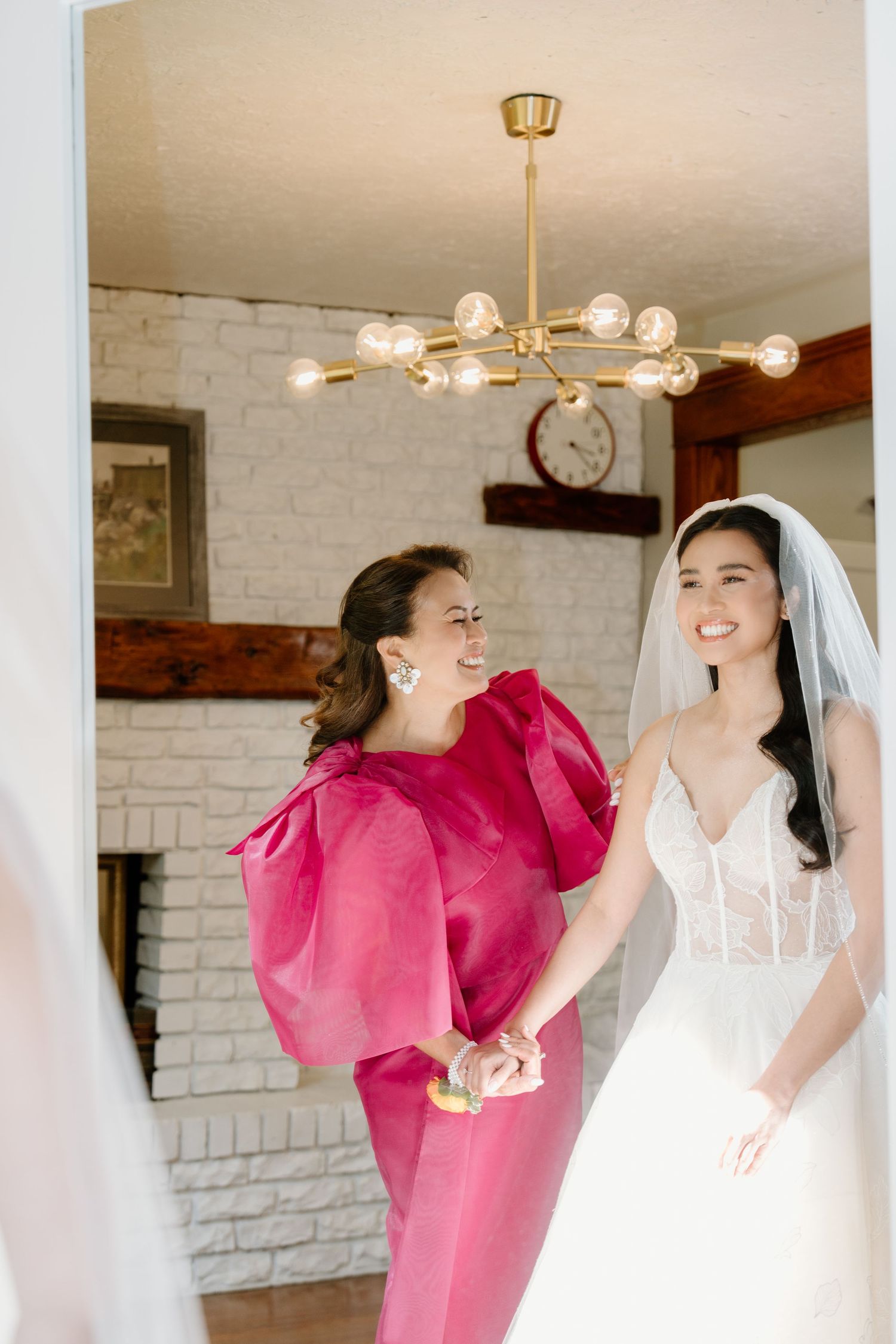 A bridesmaid in a vibrant pink dress shares a candid laughing moment with a bride in white during wedding preparations.