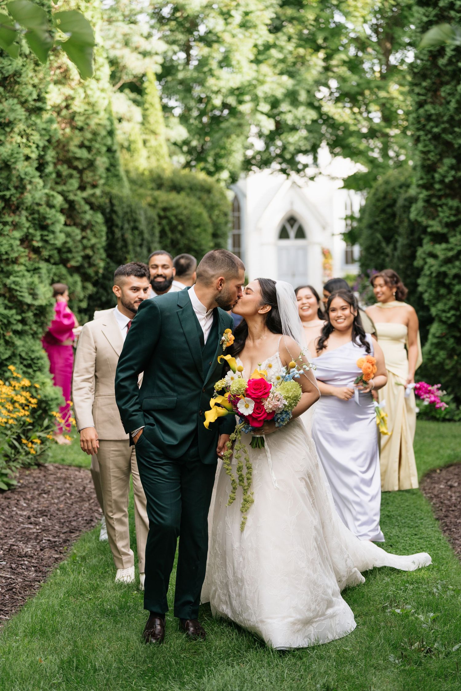A wedding party walks along a garden path with blooming flowers and greenery at an outdoor ceremony venue.