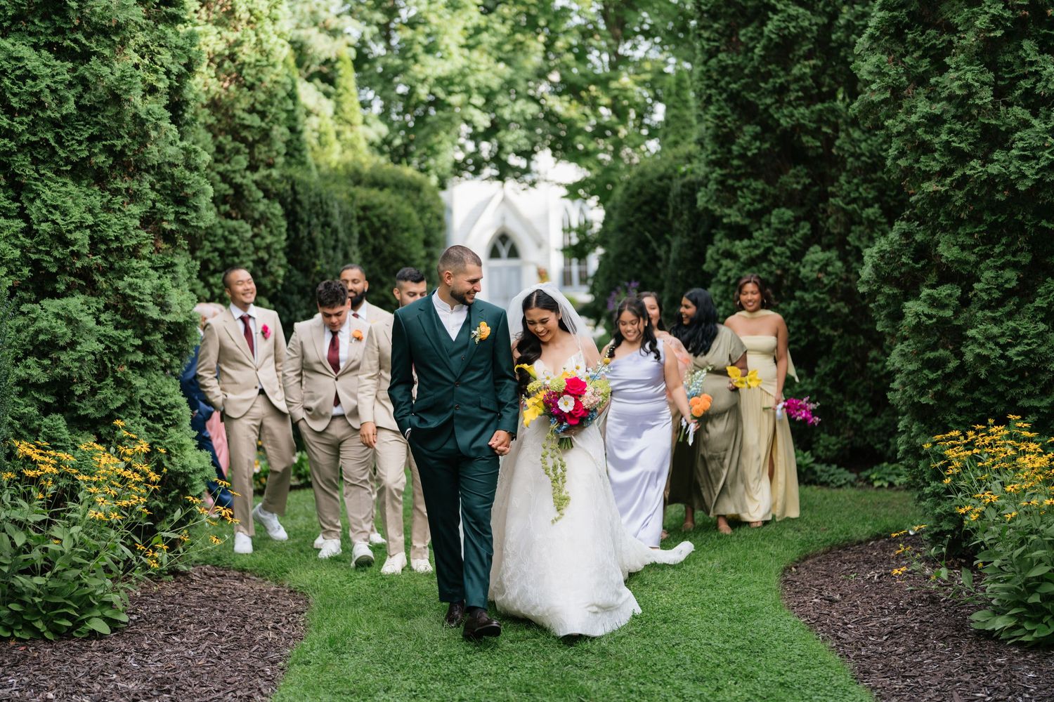 Wedding party walks along a lush garden path with tall hedges and a white chapel visible in the background.