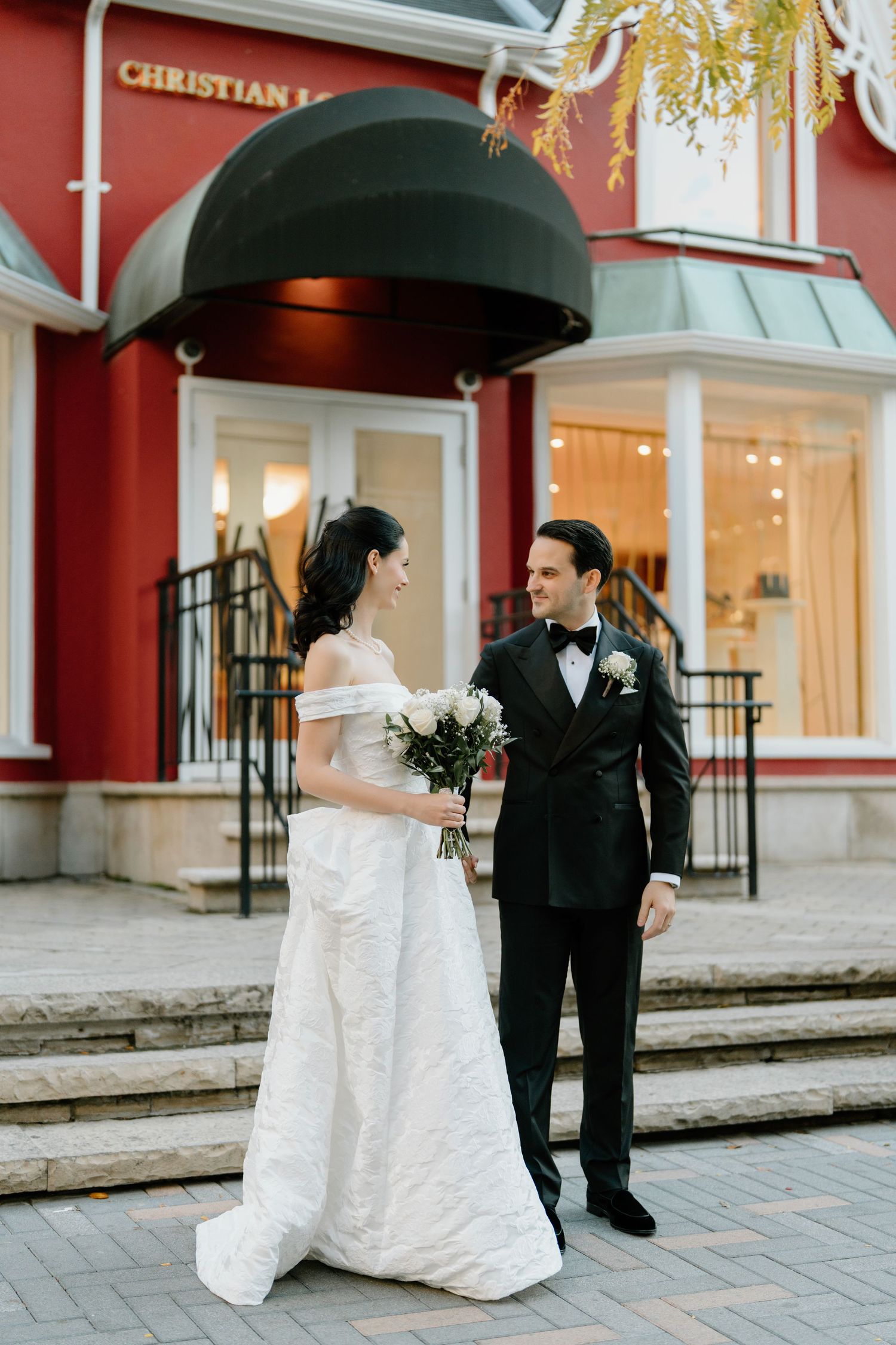 A bride and groom share an umbrella while walking along a red-walled building on a rainy wedding day.