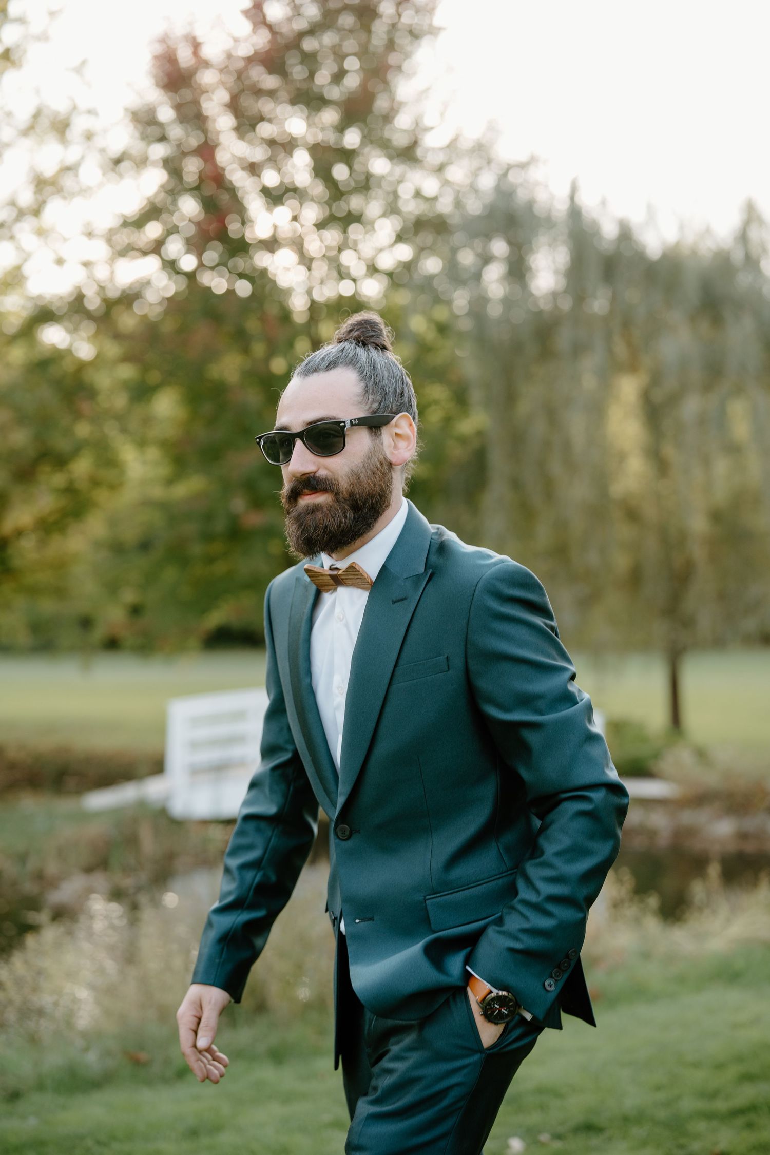 Groom in teal suit and wooden bowtie poses outdoors with sunglasses and styled hair.
