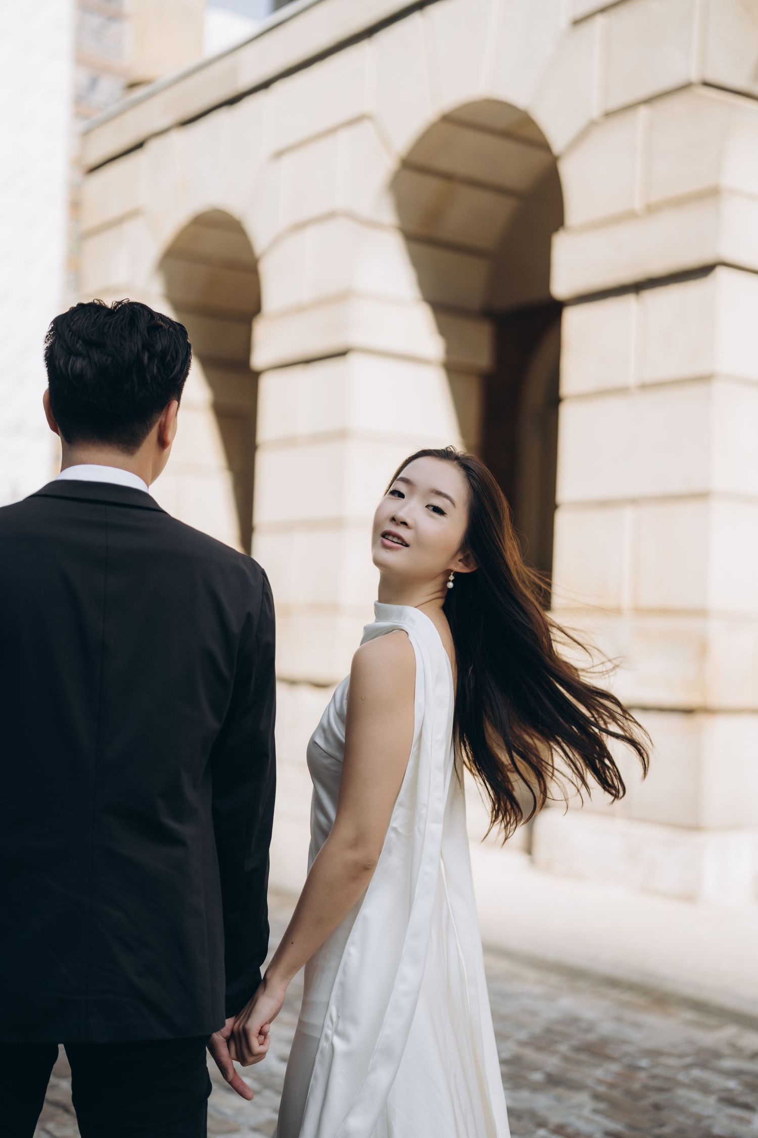 Couple in formal attire poses dramatically against stone archway building with flowing hair and romantic atmosphere.