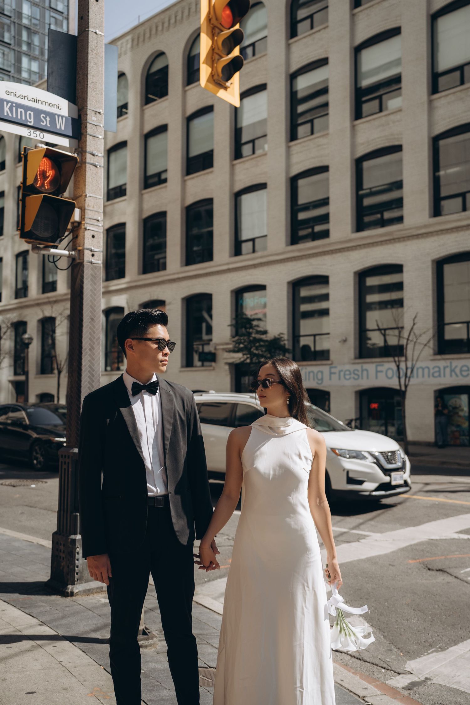 A couple in formal wedding attire stands at a city street corner with elegant buildings and traffic lights in the background.