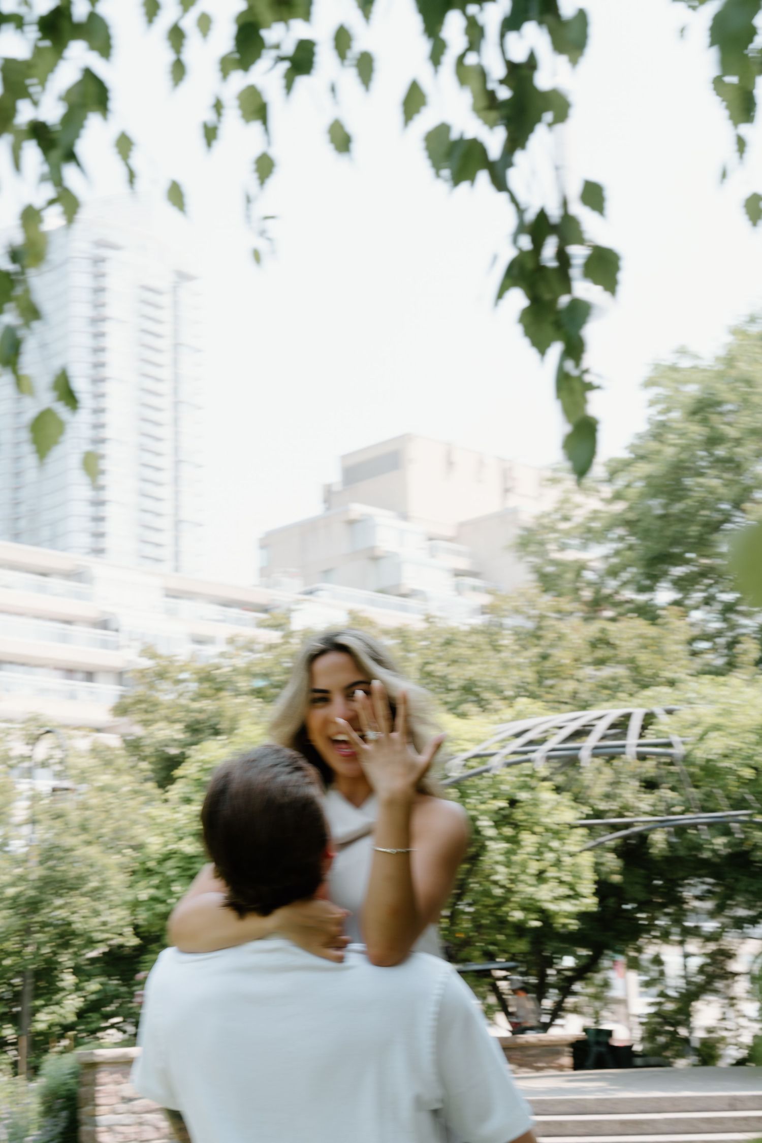 A couple shares a joyful moment outdoors with green foliage and city buildings in the background.