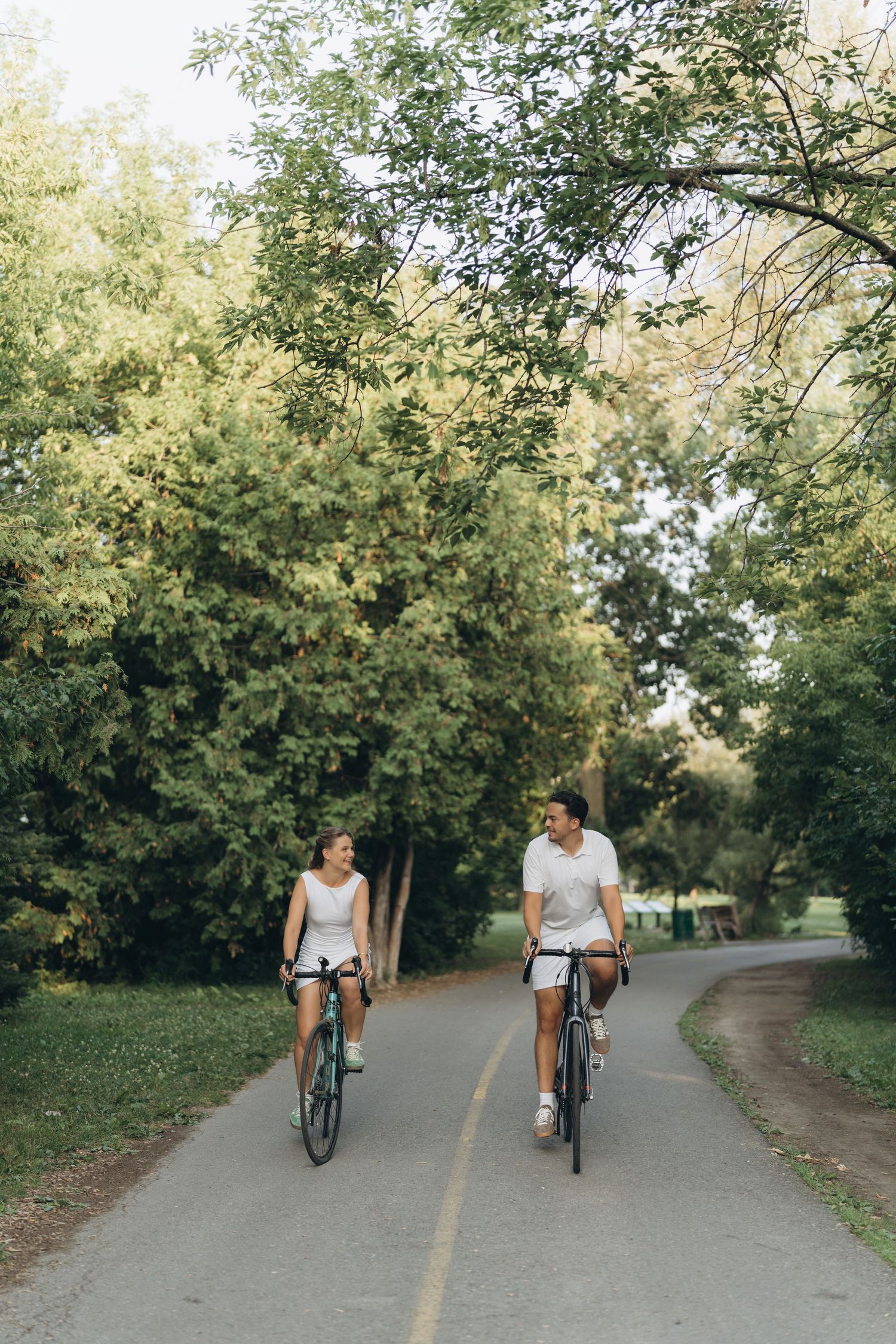 Two cyclists ride together through a tree-lined path in a park on a sunny afternoon.
